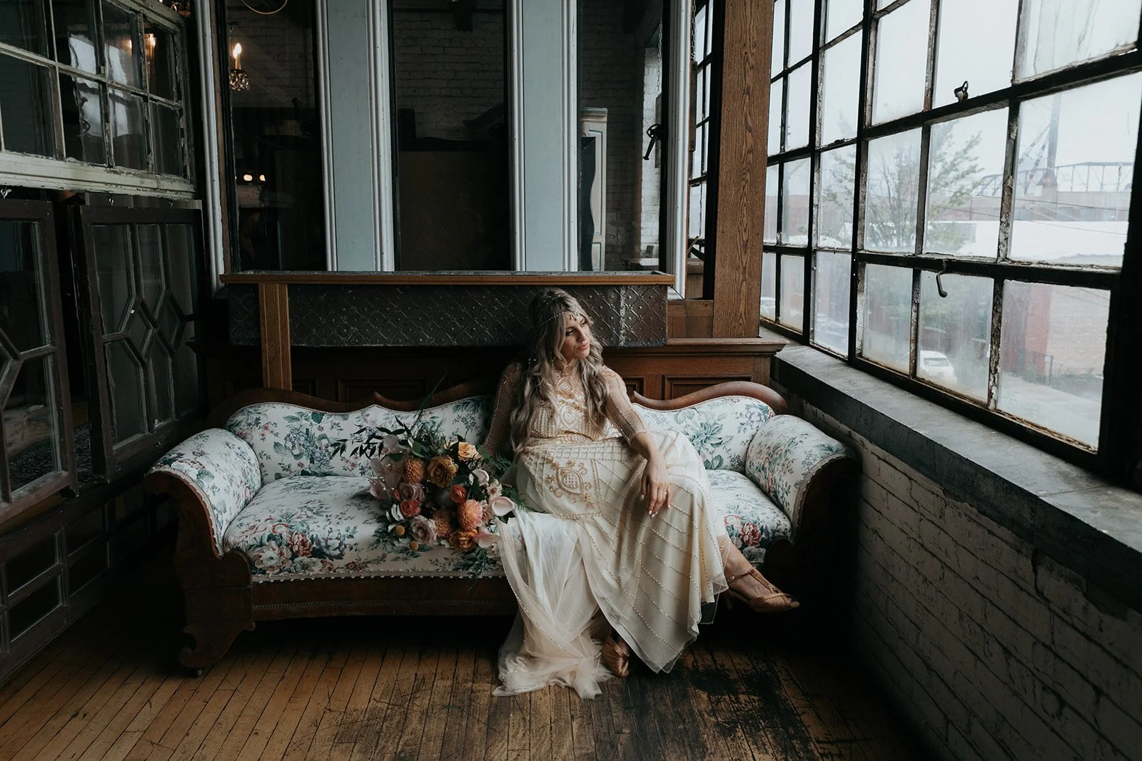 A woman in a cream-colored dress with intricate embroidery sitting on a vintage floral sofa next to a bouquet of flowers, inside a rustic-industrial room with large windows, exposed brick, and wooden accents.