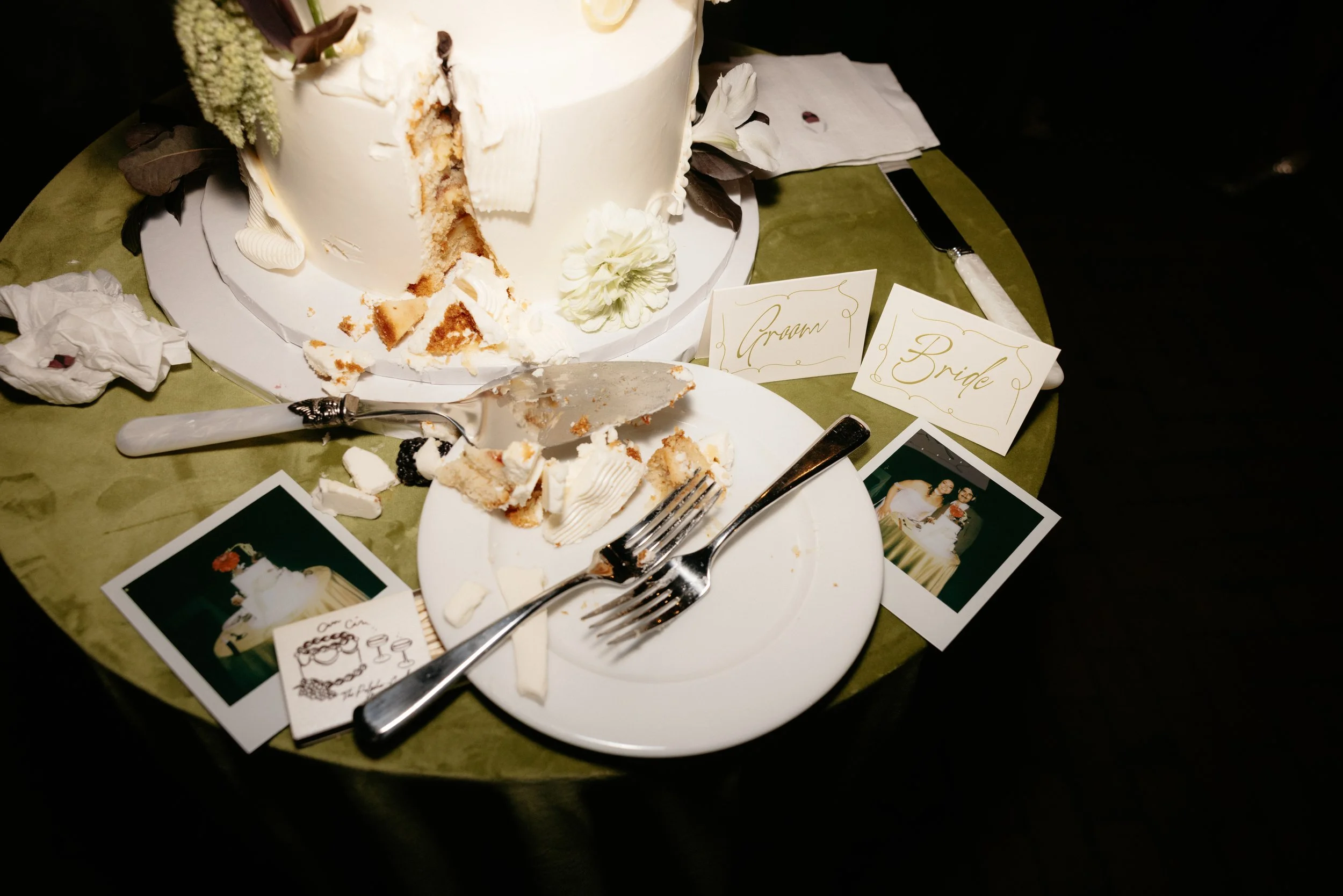 A partially eaten wedding cake on a table with wedding tags labeled Groom and Bride, along with photographs and a cake knife.