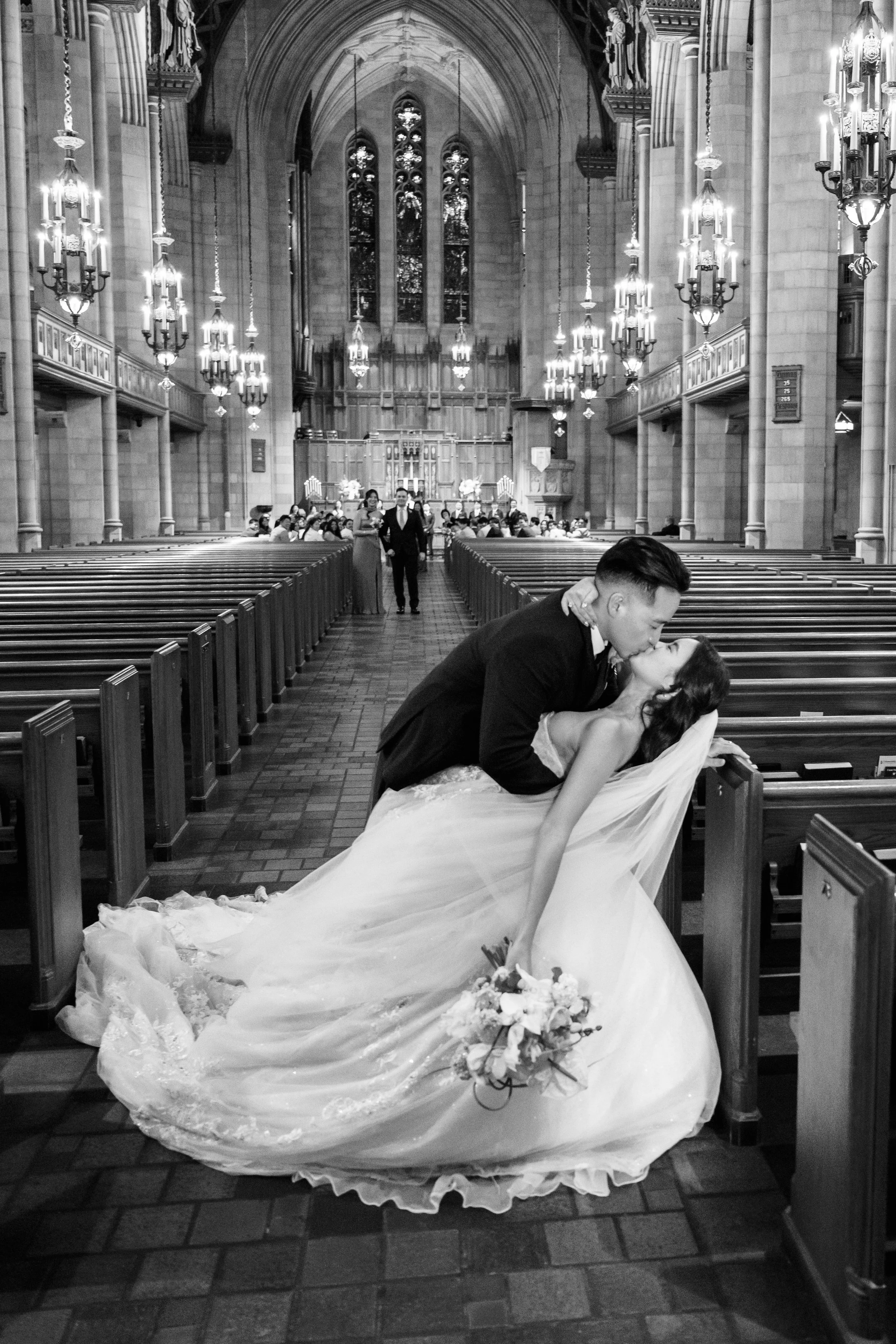A bride and groom in wedding attire kiss in a church pew, with the bride slightly leaning back and holding a bouquet. The church interior features tall stained glass windows, ornate chandeliers, and wooden pews, with guests seated in the background.