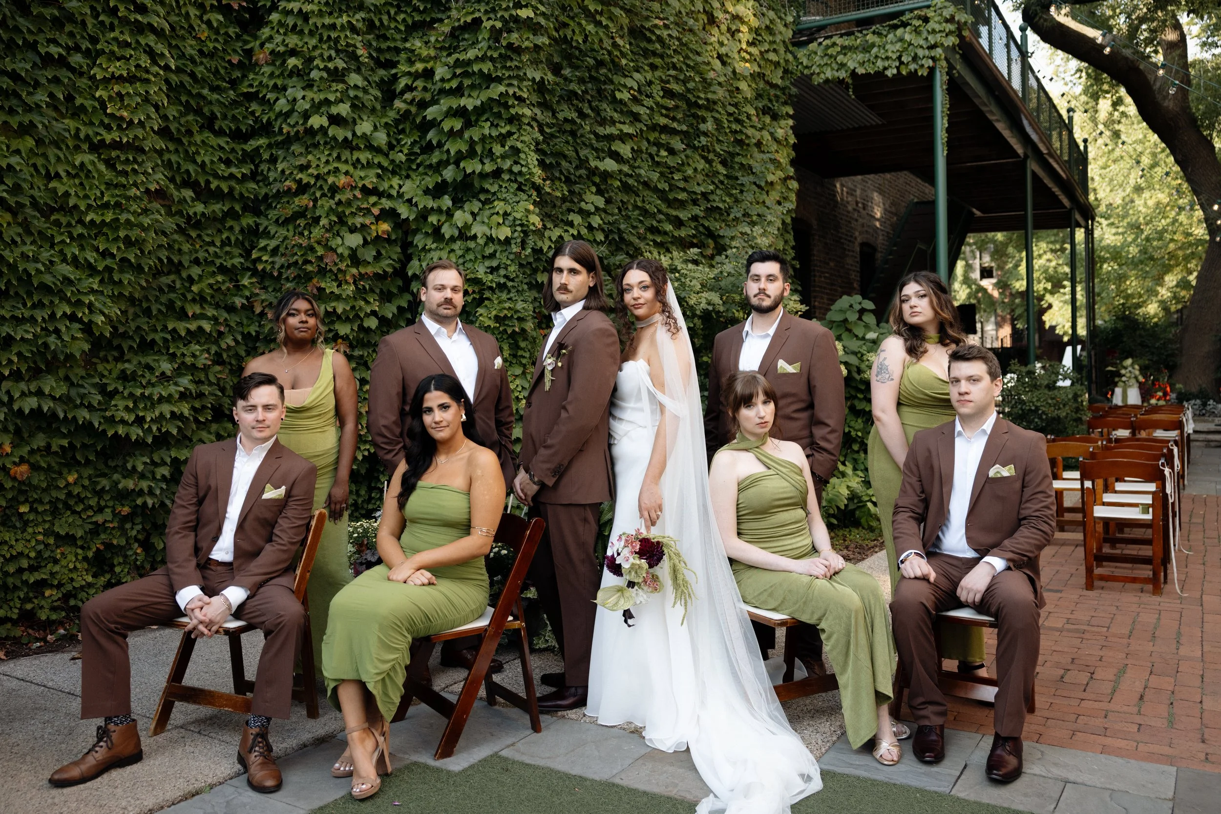 A group of people dressed for a wedding, sitting and standing outdoors in front of a green ivy-covered wall, with trees and a small outdoor staircase in the background.