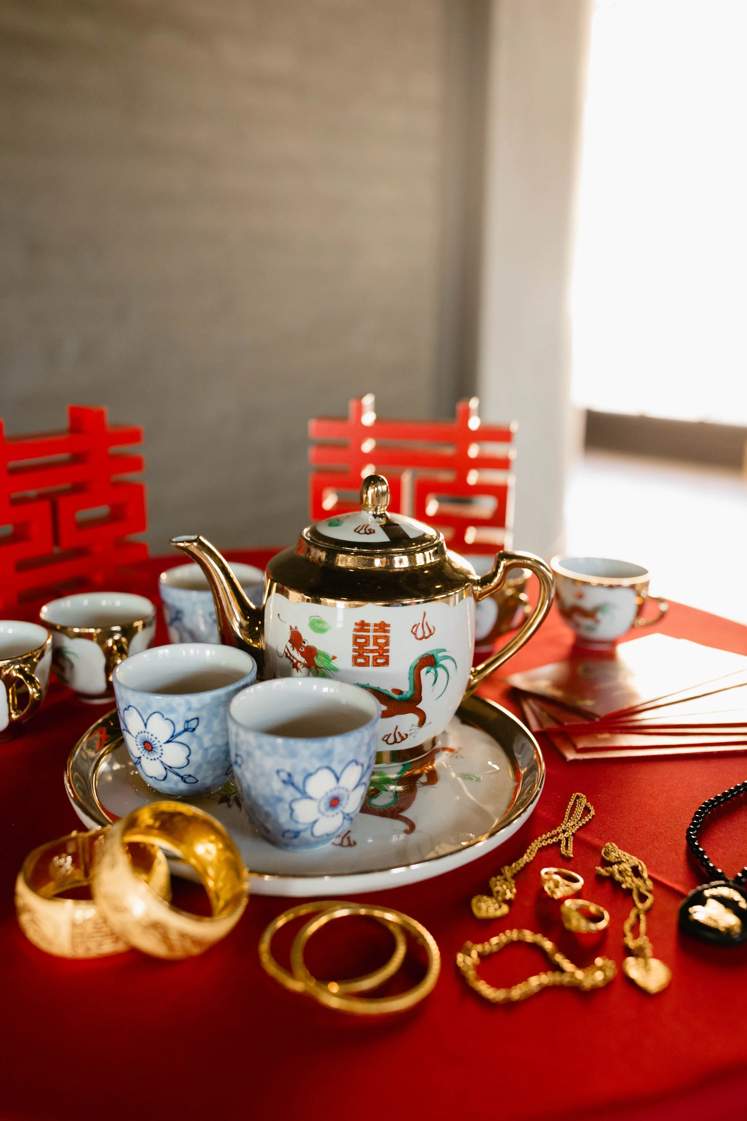 A tea set with a teapot and cups on a tray, surrounded by gold jewelry including rings, necklaces, and bracelets, displayed on a red tablecloth with red decorative screens in the background.