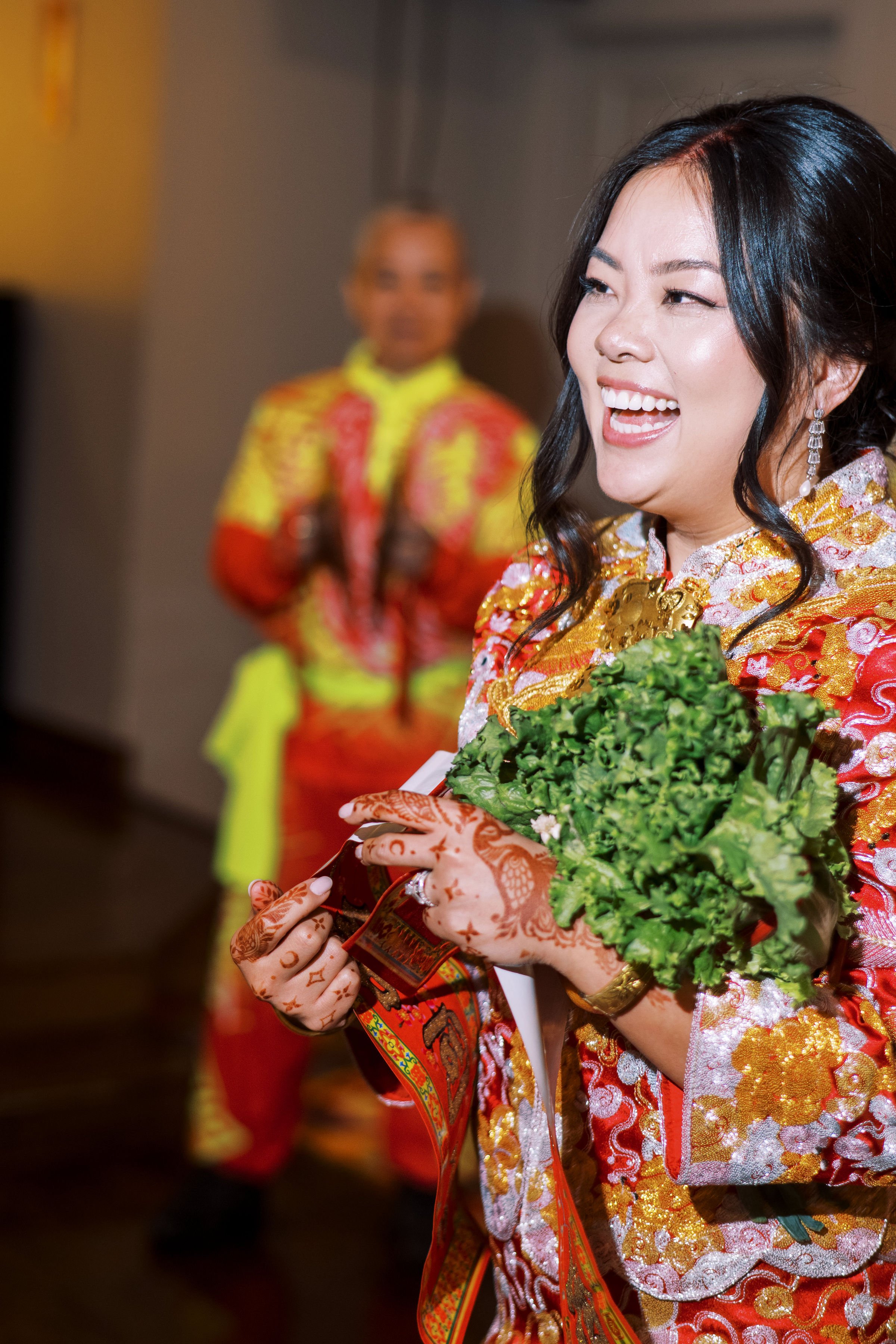 A woman dressed in a colorful traditional Asian outfit holding a bouquet of green leafy vegetables, with another person in a similar outfit blurred in the background.