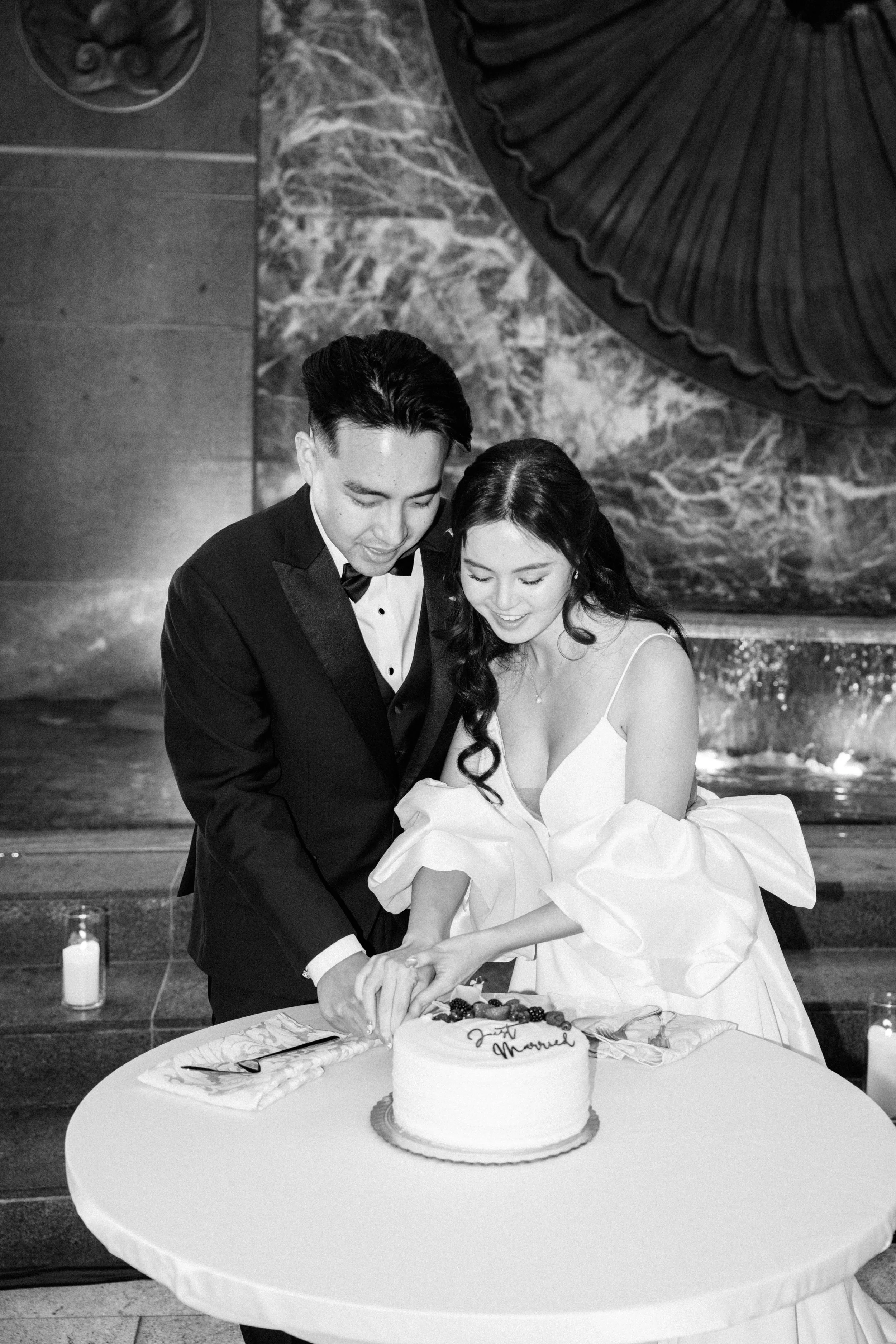 A newlywed couple cutting their wedding cake at a reception, with the cake reading 'Just Married,' surrounded by candles and a large decorative umbrella overhead.