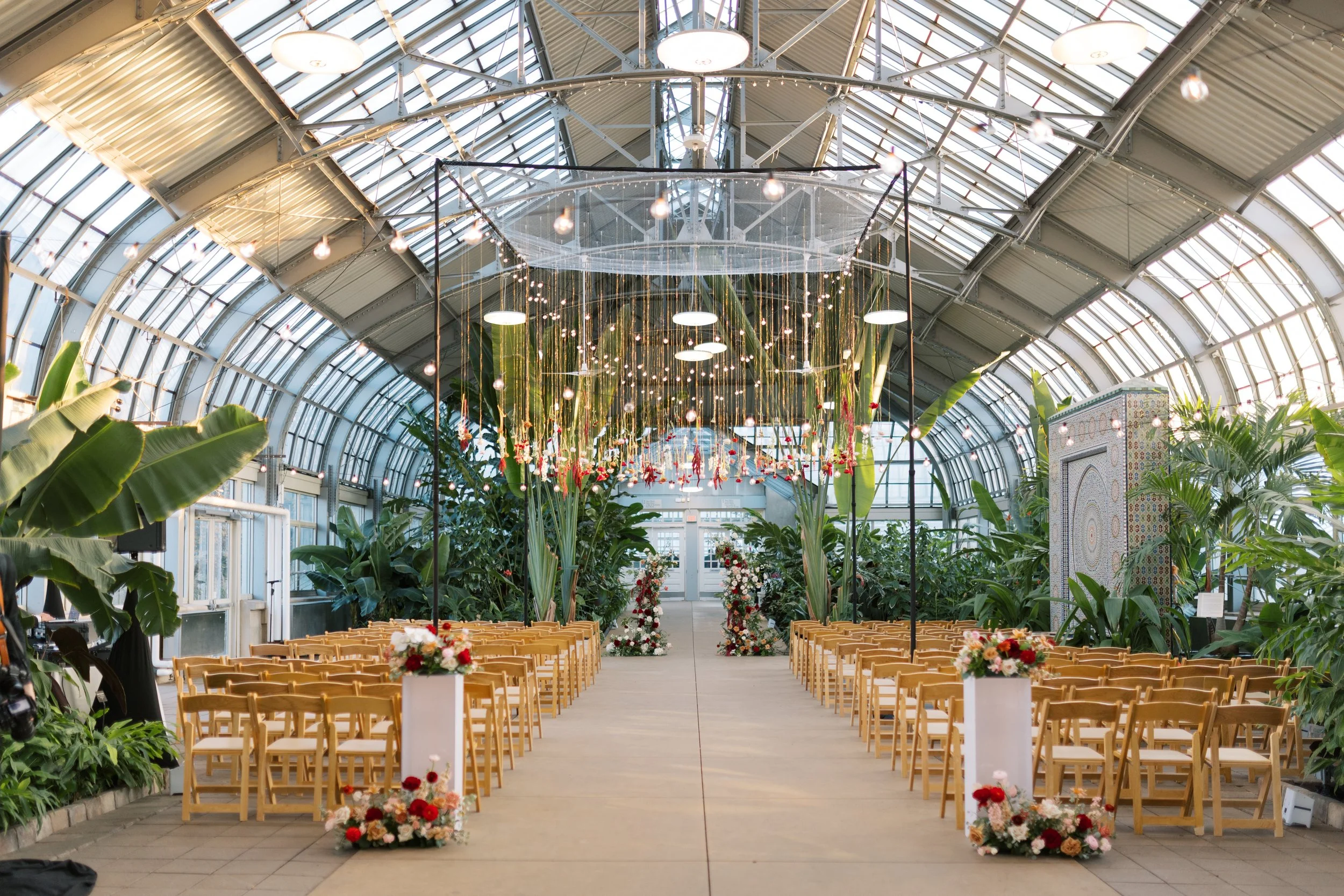 An indoor botanical garden or greenhouse decorated for a wedding ceremony with rows of wooden chairs on either side of a central aisle, floral arrangements, and hanging string lights beneath a glass arched ceiling.