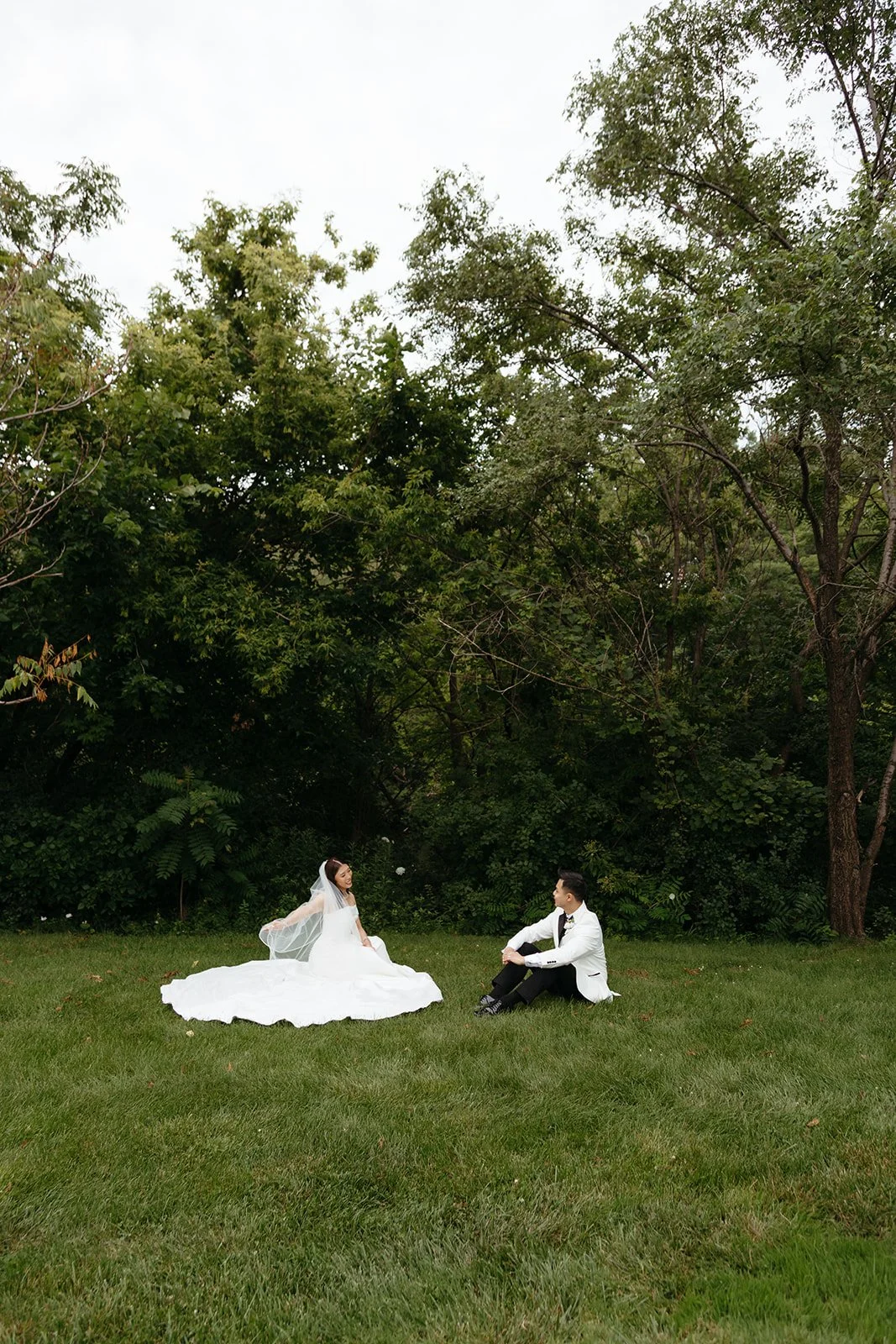 A bride in a white wedding gown and veil is sitting on the grass, smiling, while a groom in a white tuxedo with black pants and bowtie is sitting nearby, in front of a background of tall green trees and bushes.