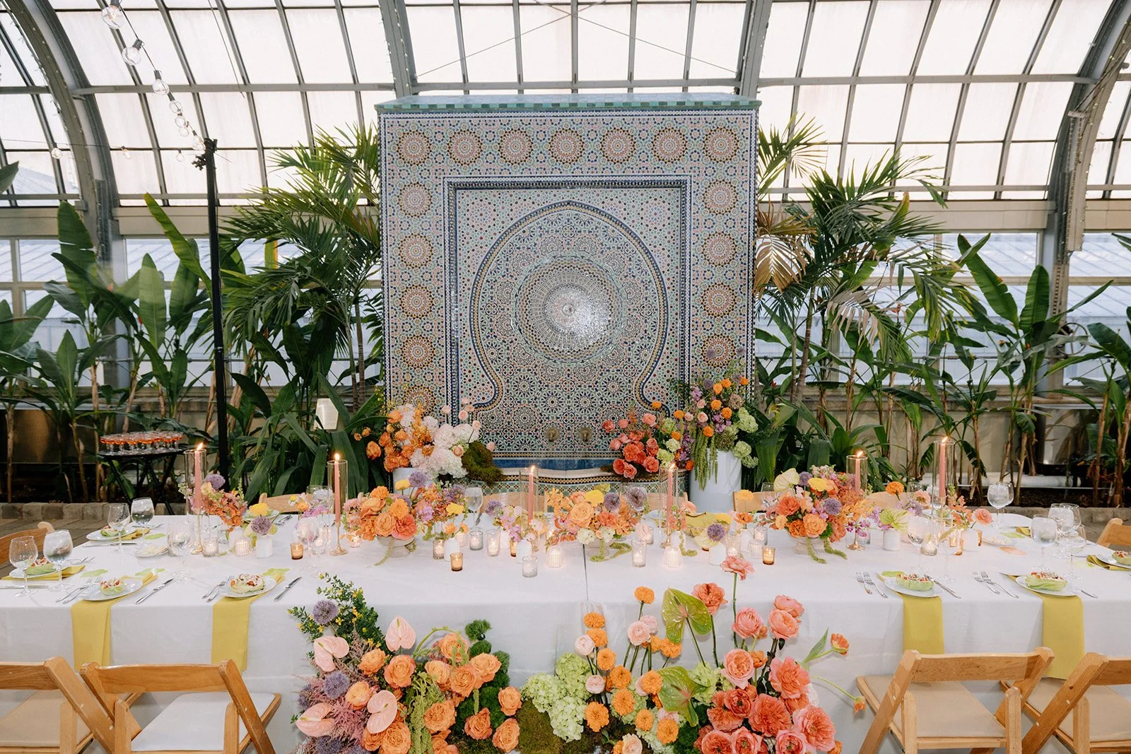 A beautifully decorated banquet table with floral arrangements, candles, and glassware, set against a backdrop of intricate mosaic art and surrounded by lush green plants in an enclosed glass structure.