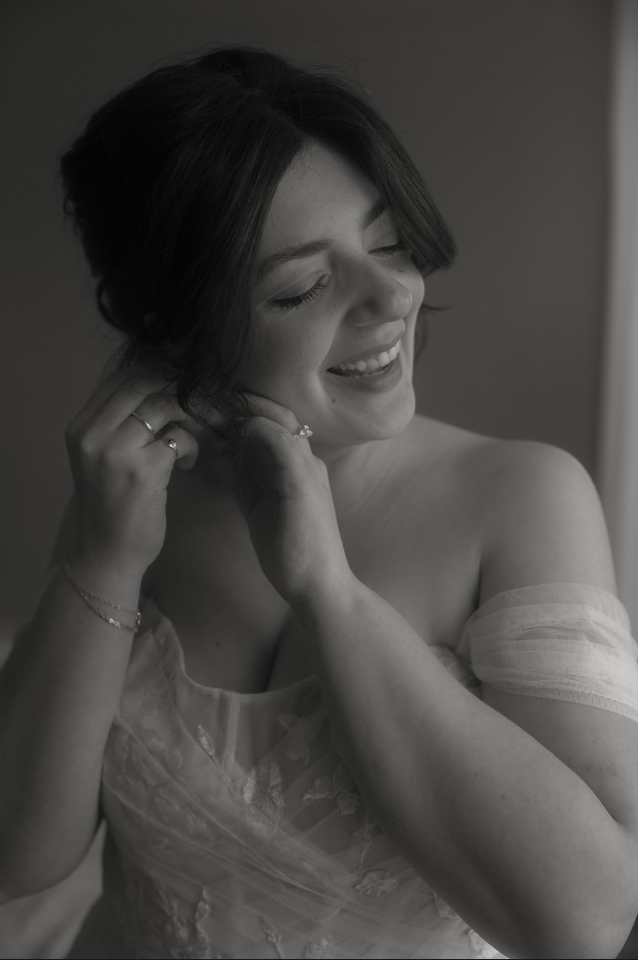 A woman in a dress smiling while adjusting her earring with her eyes closed, in black and white.