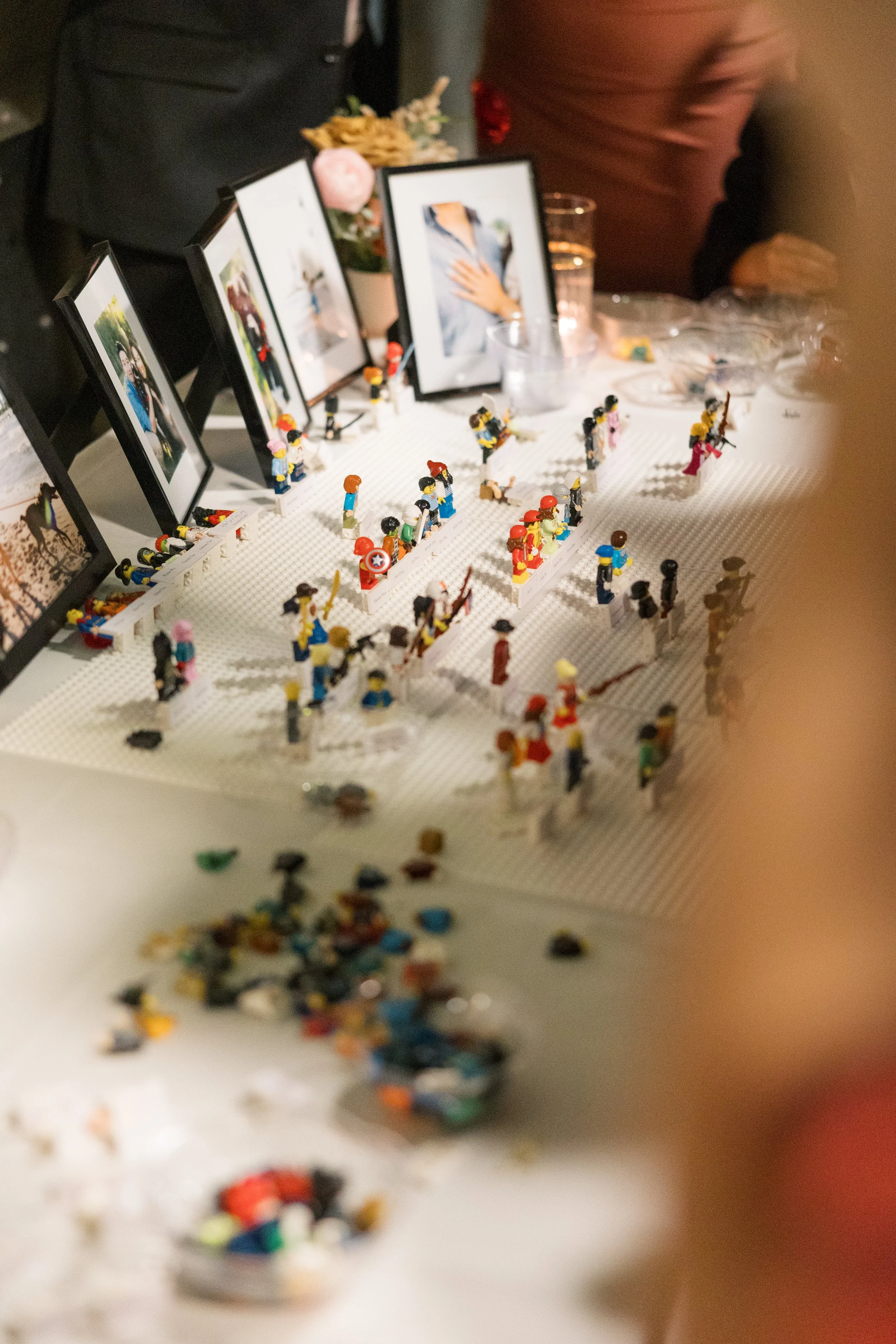 A collection of LEGO minifigures on a white baseplate, with framed photographs, a purple flower, and a black bag in the background.