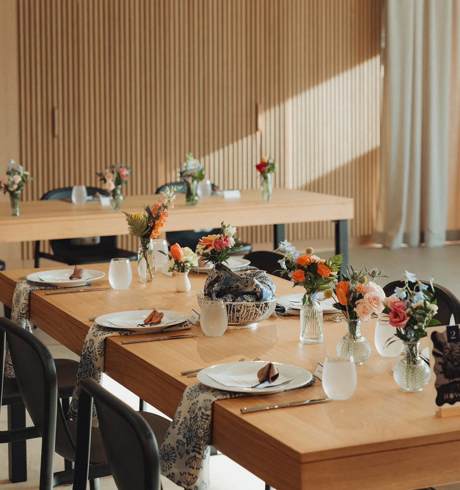A set dining table decorated with floral arrangements in vases, decorated plates, utensils, napkins, and glasses in a room with wooden wall paneling and sunlight streaming through a curtain.