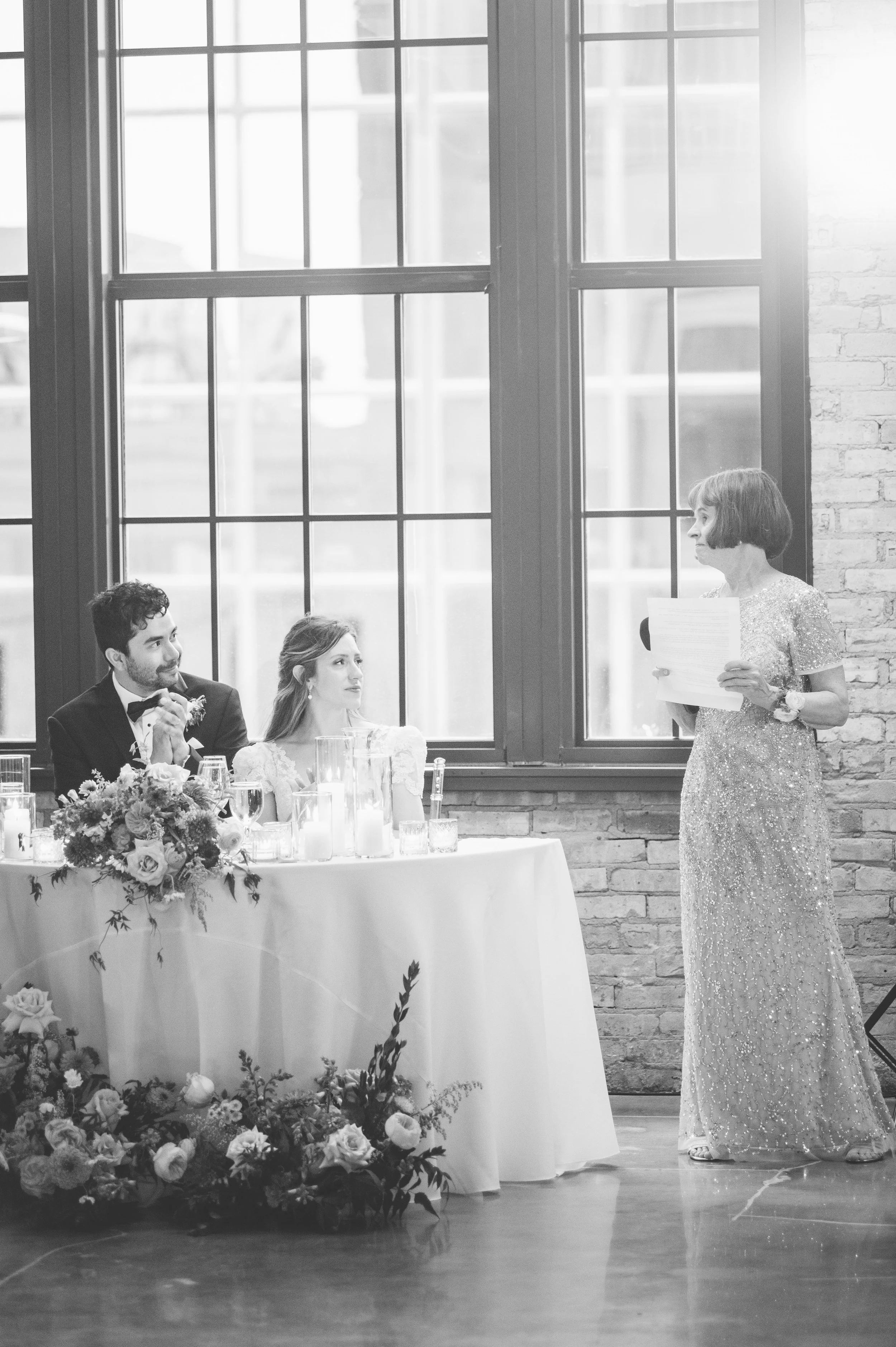 A woman giving a speech at a wedding reception, standing next to a table with floral decorations, while three people, a man and two women, are seated at the table listening. Large windowpanes and a brick wall are in the background.
