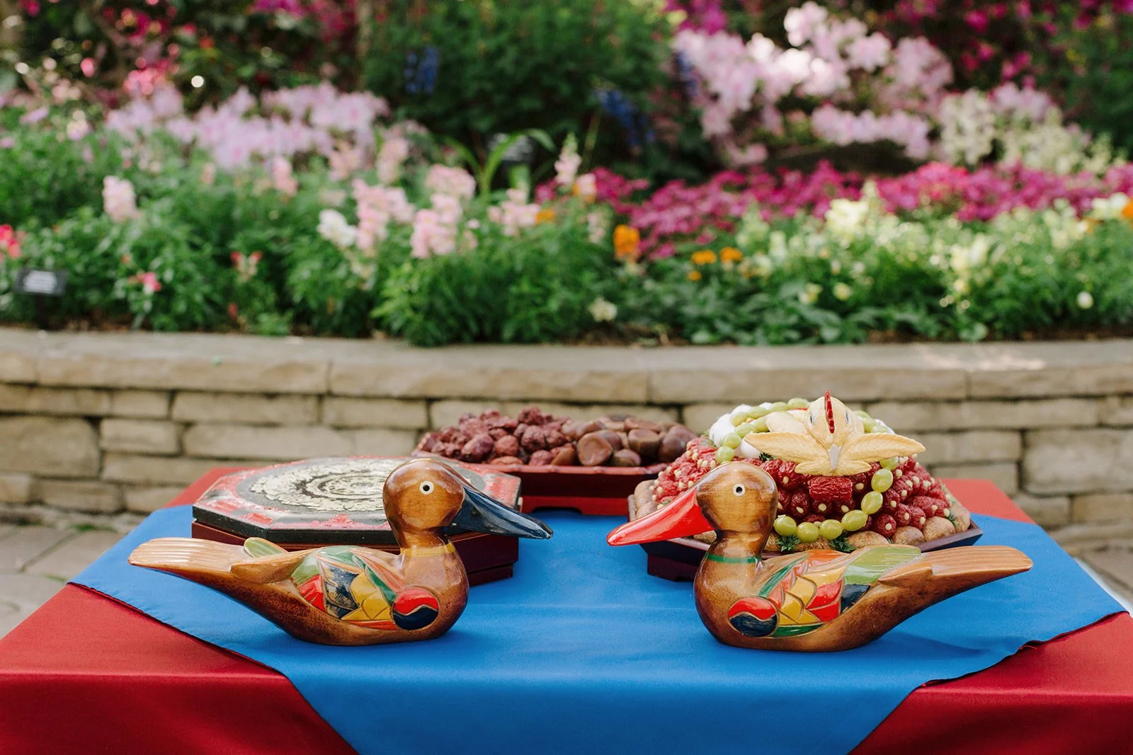 Decorative wooden duck figurines on a table with a red and blue tablecloth, with a colorful fruit arrangement and a bowl of nuts in the background, outdoors with blooming flowers and a brick wall.