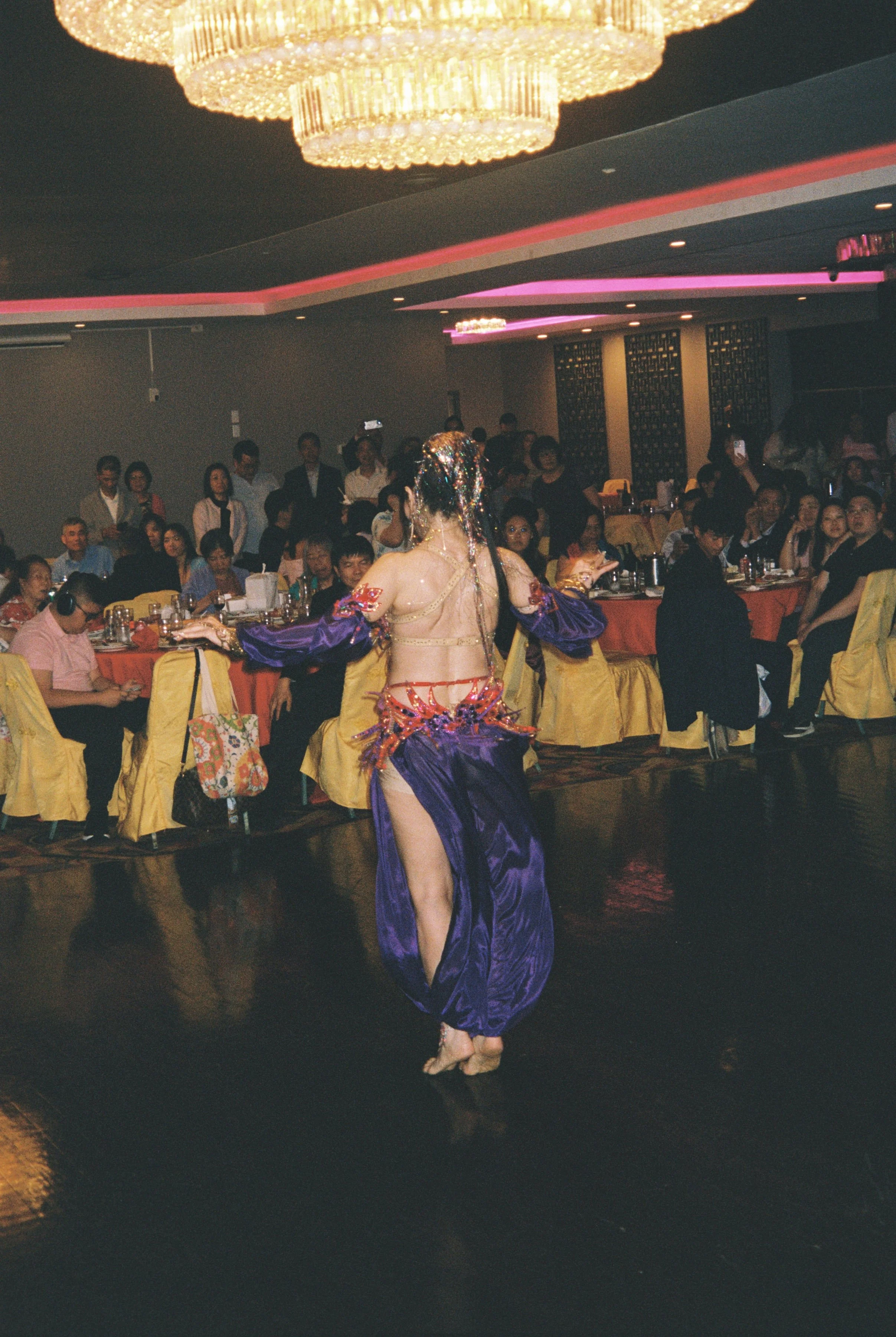 A woman performs a dance in a banquet hall with seated and standing guests watching.