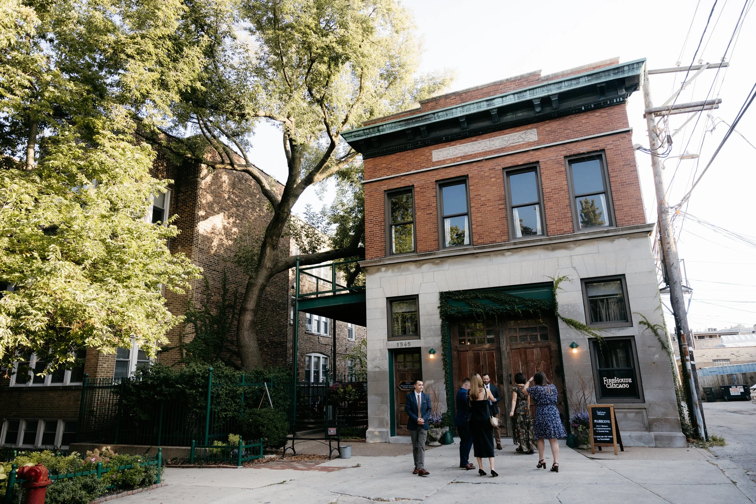 People in formal attire gathered outside a brick building labeled 'Firehouse Chicago' with a sign for parking nearby, surrounded by trees and urban infrastructure.