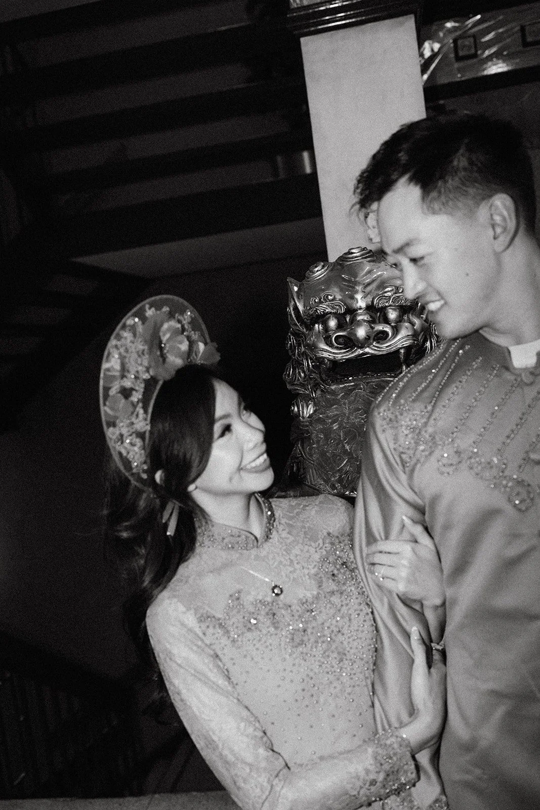 A smiling woman wearing a traditional Vietnamese dress and a large decorative hat looks at a man in a traditional dress, with a lion mask in the background.