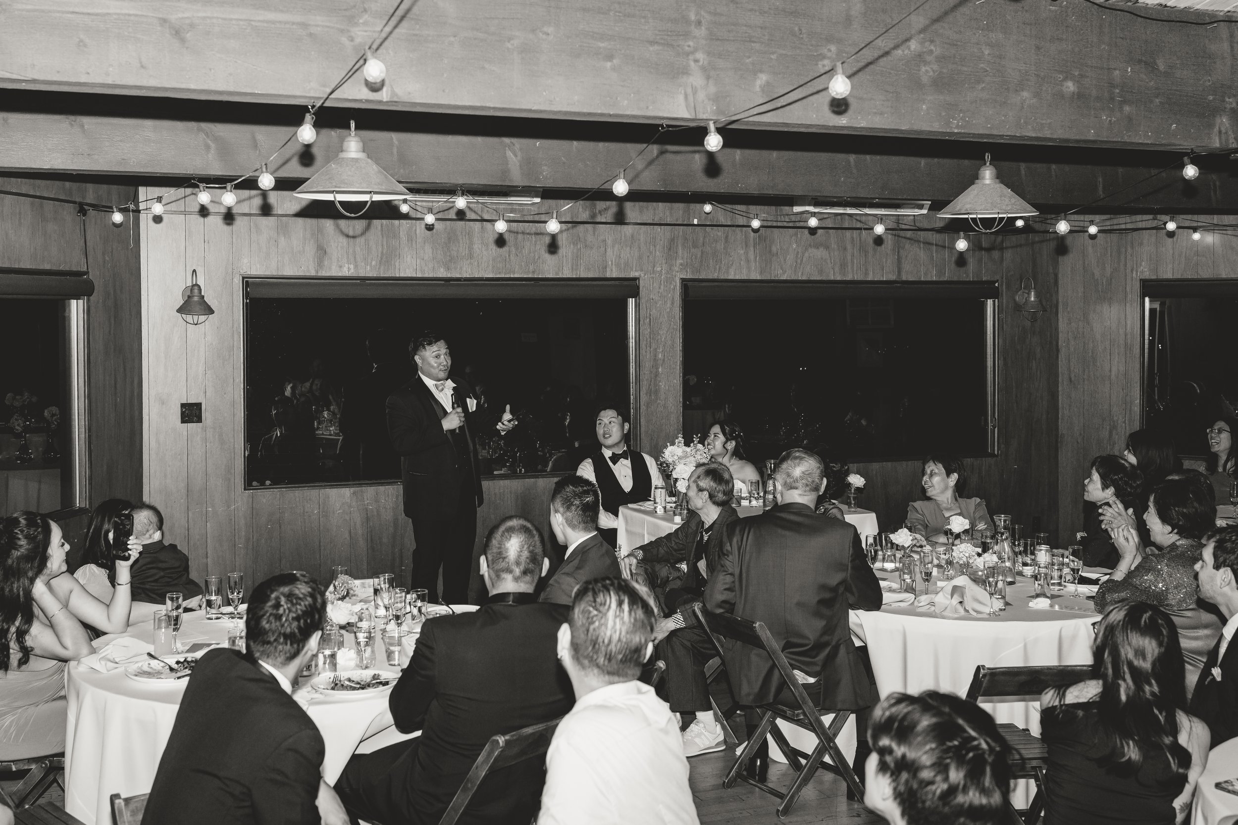 A black and white photo of a formal dinner event with guests sitting at tables, a man in a tuxedo giving a toast or speech, and a woman in formal wear sitting nearby, with decorative string lights hanging from the ceiling.