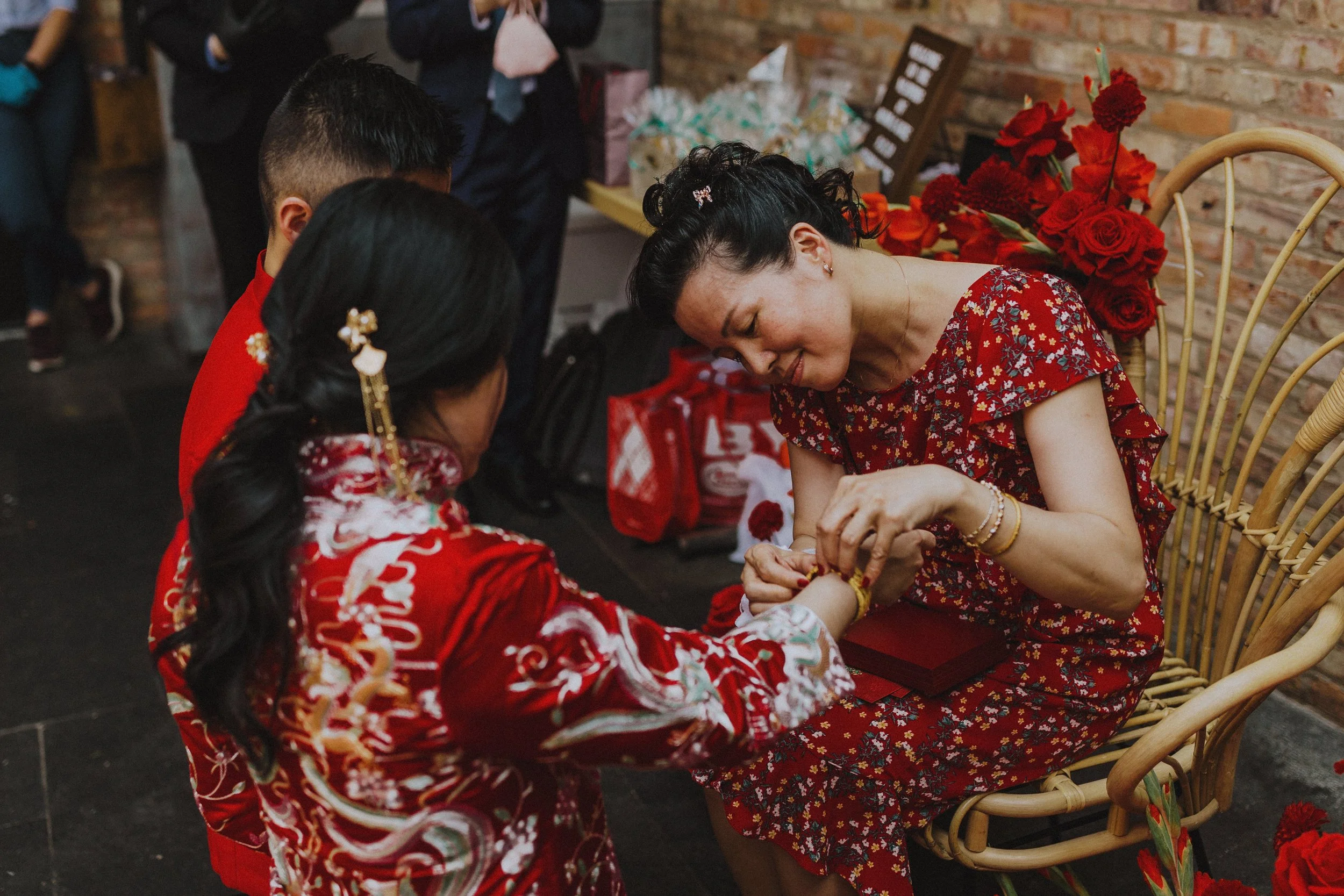 A woman in a red floral dress is sitting and getting a henna tattoo on her hand by a girl dressed in a traditional red and white embroidered dress. The woman is smiling with her eyes closed, and there are red flowers in the background.