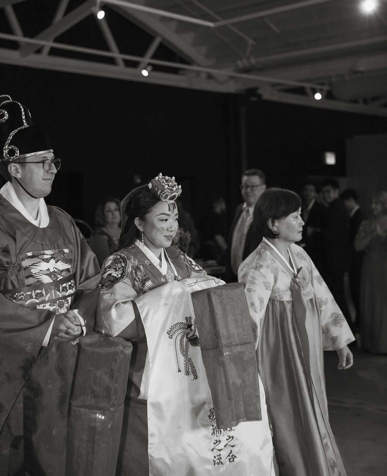 Three individuals dressed in traditional Asian attire, possibly during a cultural ceremony or event, in an indoor setting with other people in the background.
