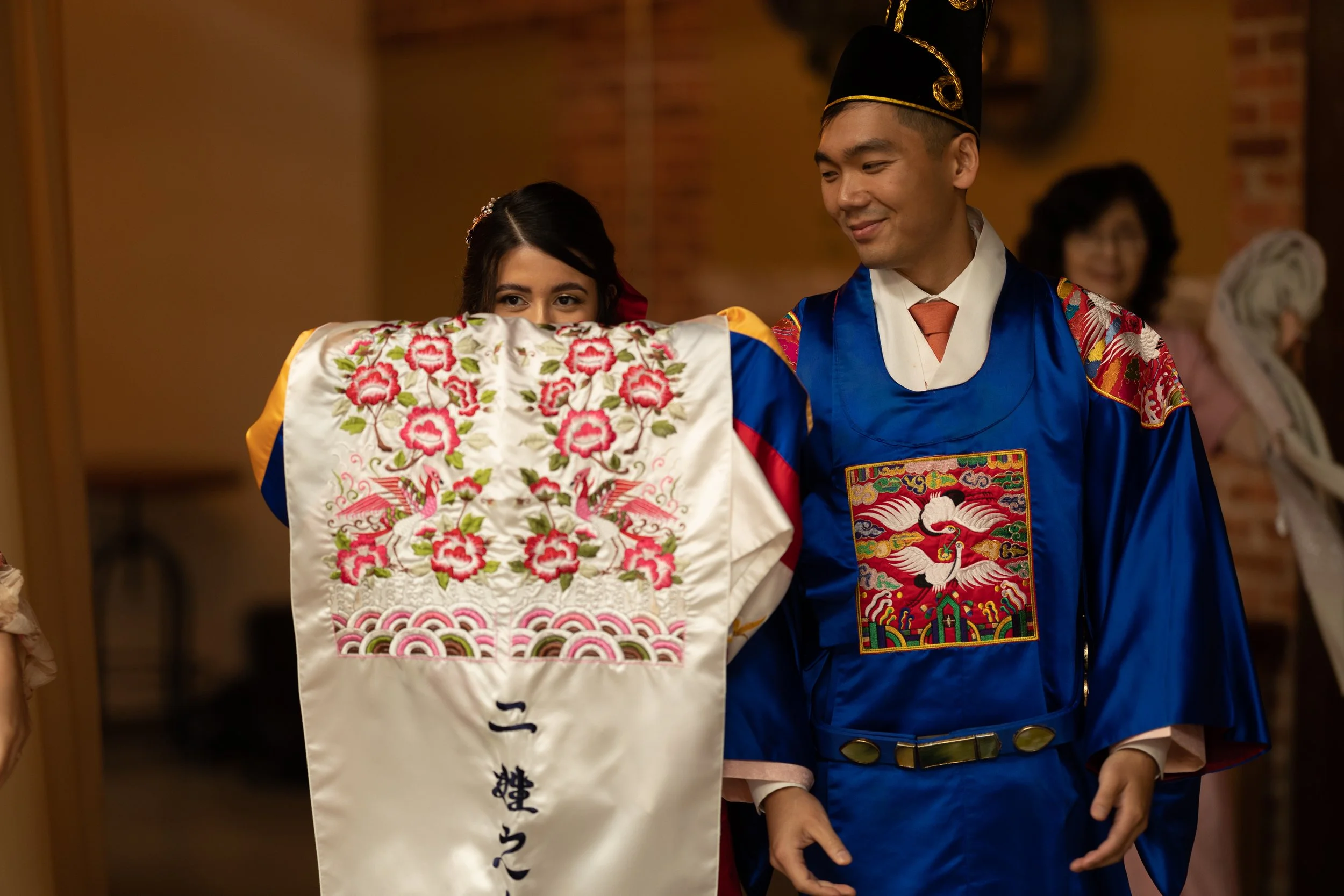 A young woman and a man dressed in traditional Korean wedding attire are participating in a ceremony; the woman holds a embroidered silk pillow with floral designs, and the man smiles at her.