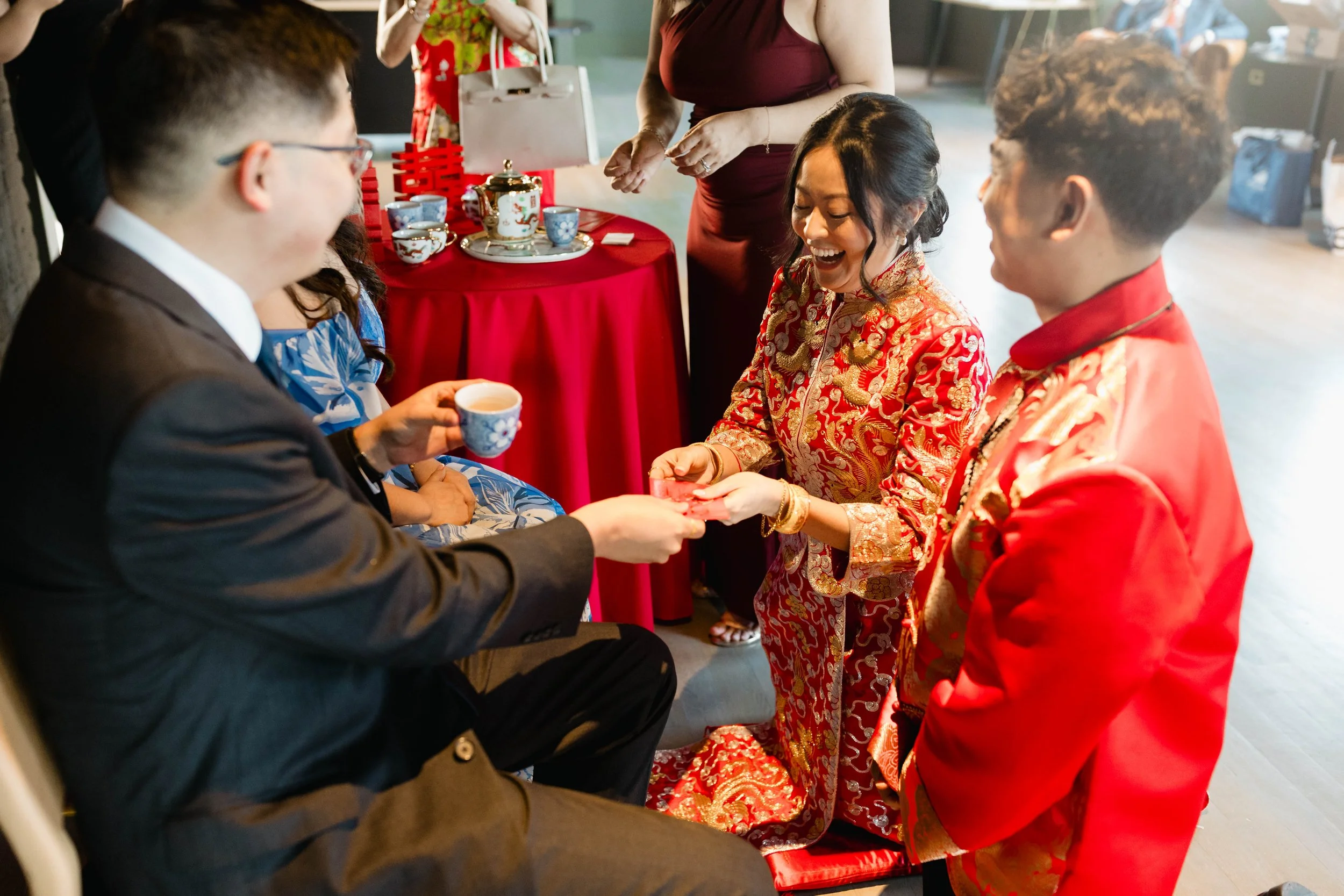 People at a traditional Chinese wedding ceremony, with the bride in a red and gold gown and the groom in a suit, exchanging rings in front of friends and family.