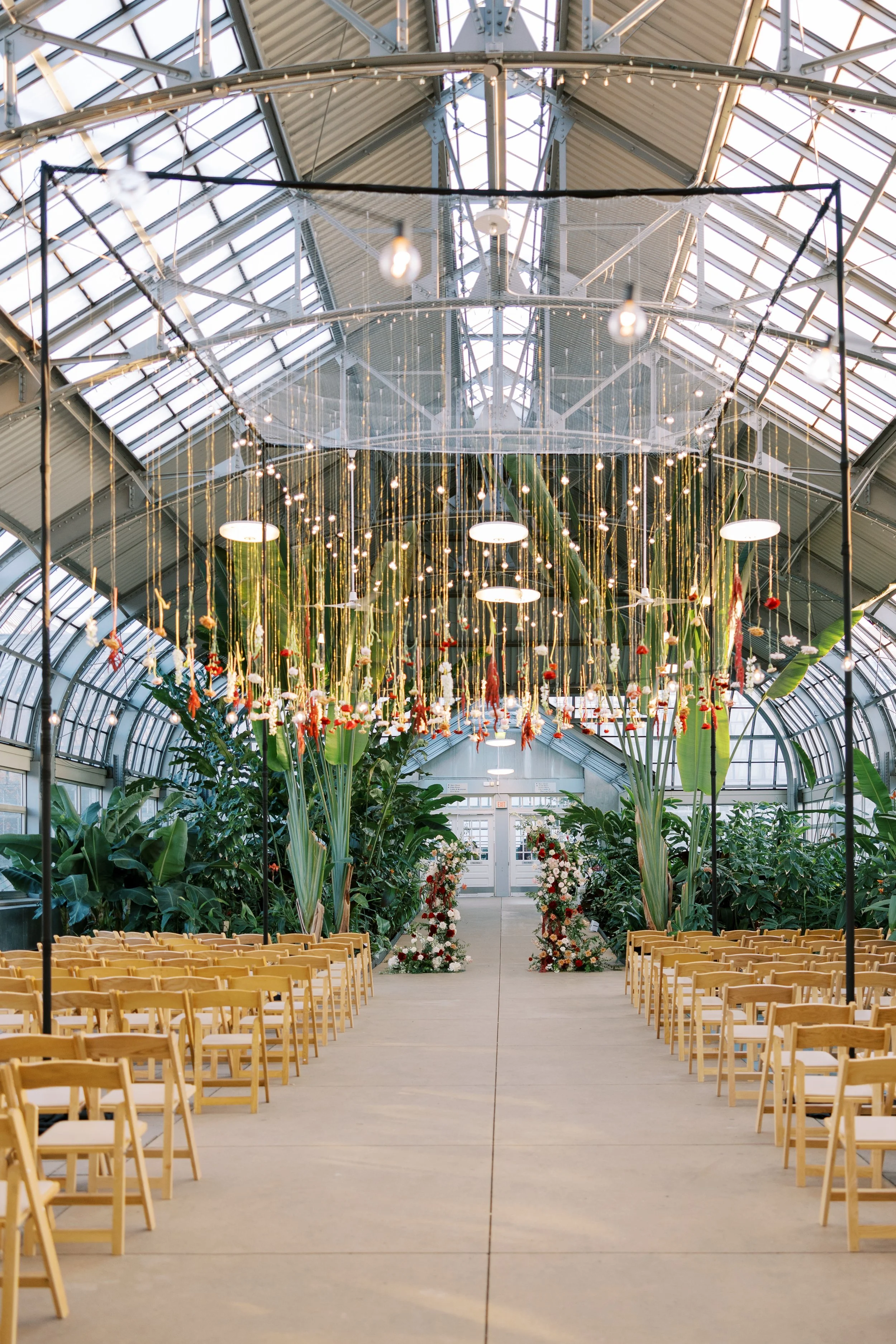 Indoor greenhouse decorated for a wedding with roses and greenery along the aisle, rows of wooden chairs on either side, hanging lights, and a glass ceiling.