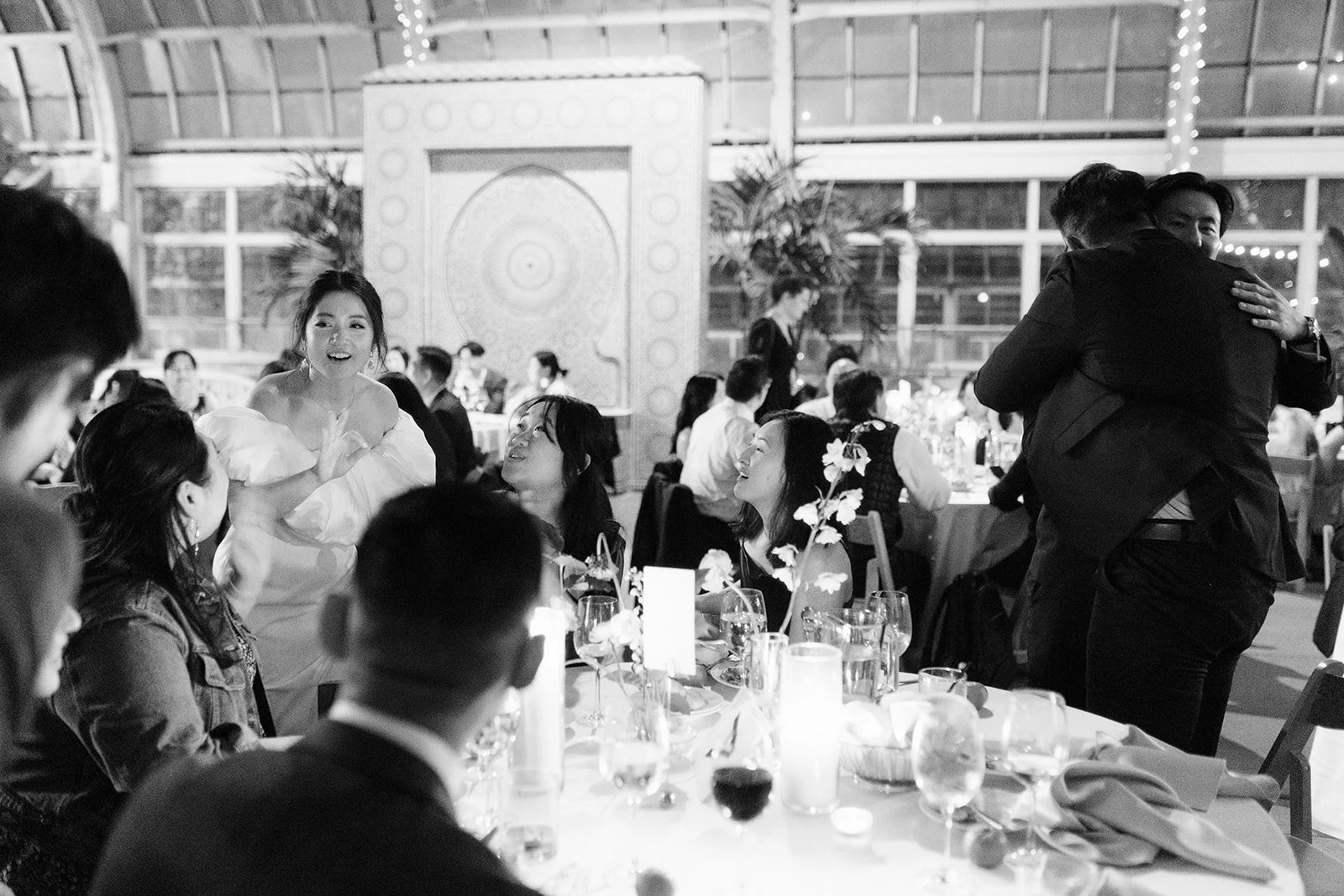 People at a formal event, some sitting at tables and others standing and hugging, with a woman in a white dress speaking to guests in a decorated venue.