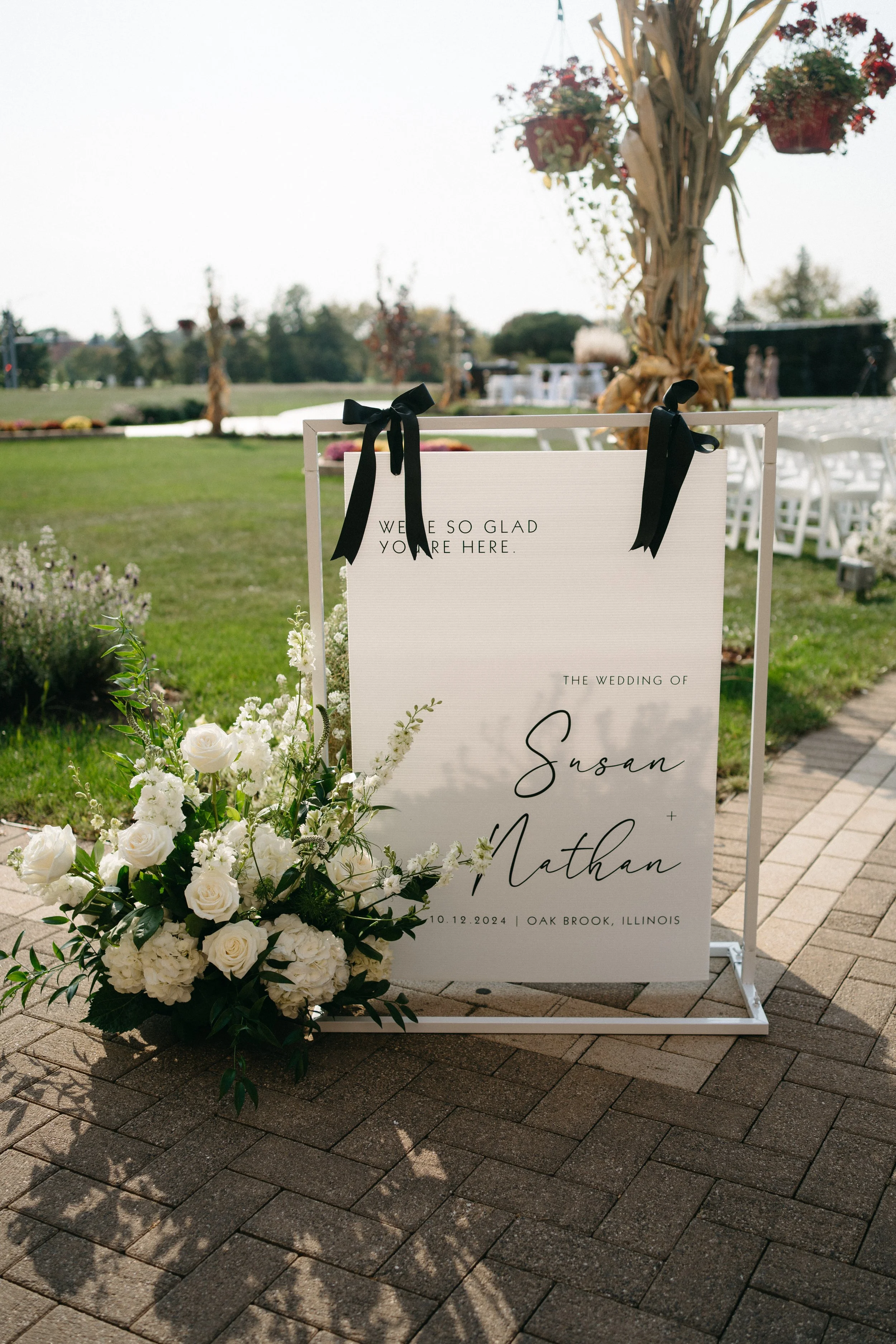 Wedding sign with white floral arrangement at Oak Brook, Illinois, on October 12, 2024, featuring black ribbons and outdoor setting.