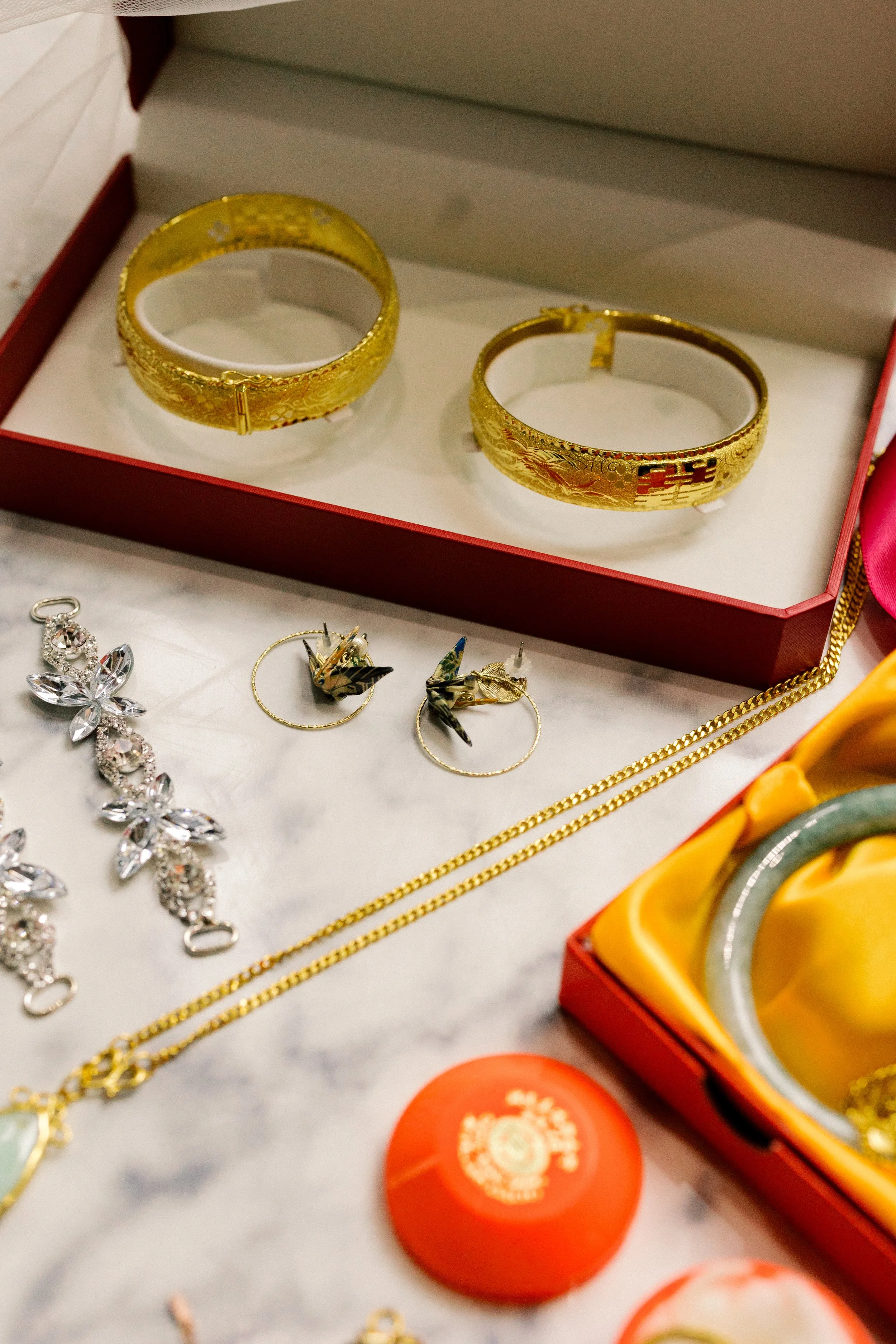 Gold jewelry, including two wide gold bracelets with intricate designs, a pair of gold earrings with bird motifs, a silver-colored bracelet with clear stones, and gold chains, displayed in a red jewelry box and on a white surface.