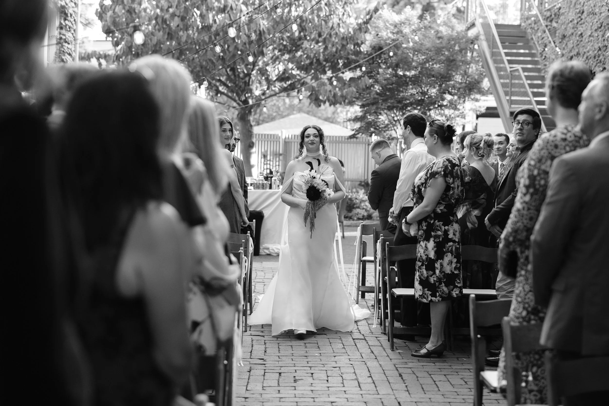 A wedding ceremony outdoors with a bride in a white gown holding a bouquet, standing among guests seated in chairs, with string lights and trees in the background.
