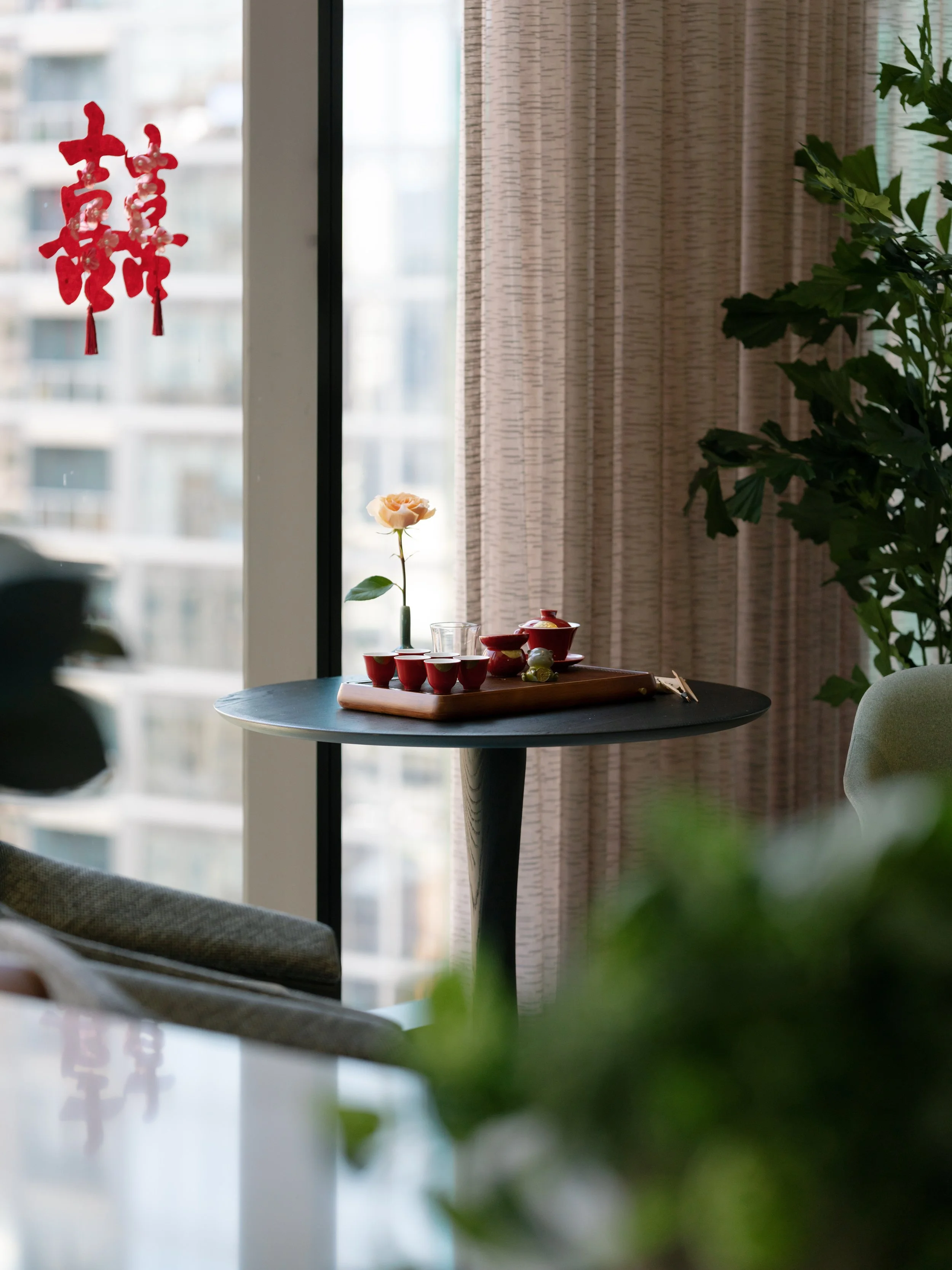A small round table with a wooden tray holding a vase with a single peach-colored flower, a tea set with red cups, and some decorative stones. In the background, there is a window, beige curtains, and some indoor plants.