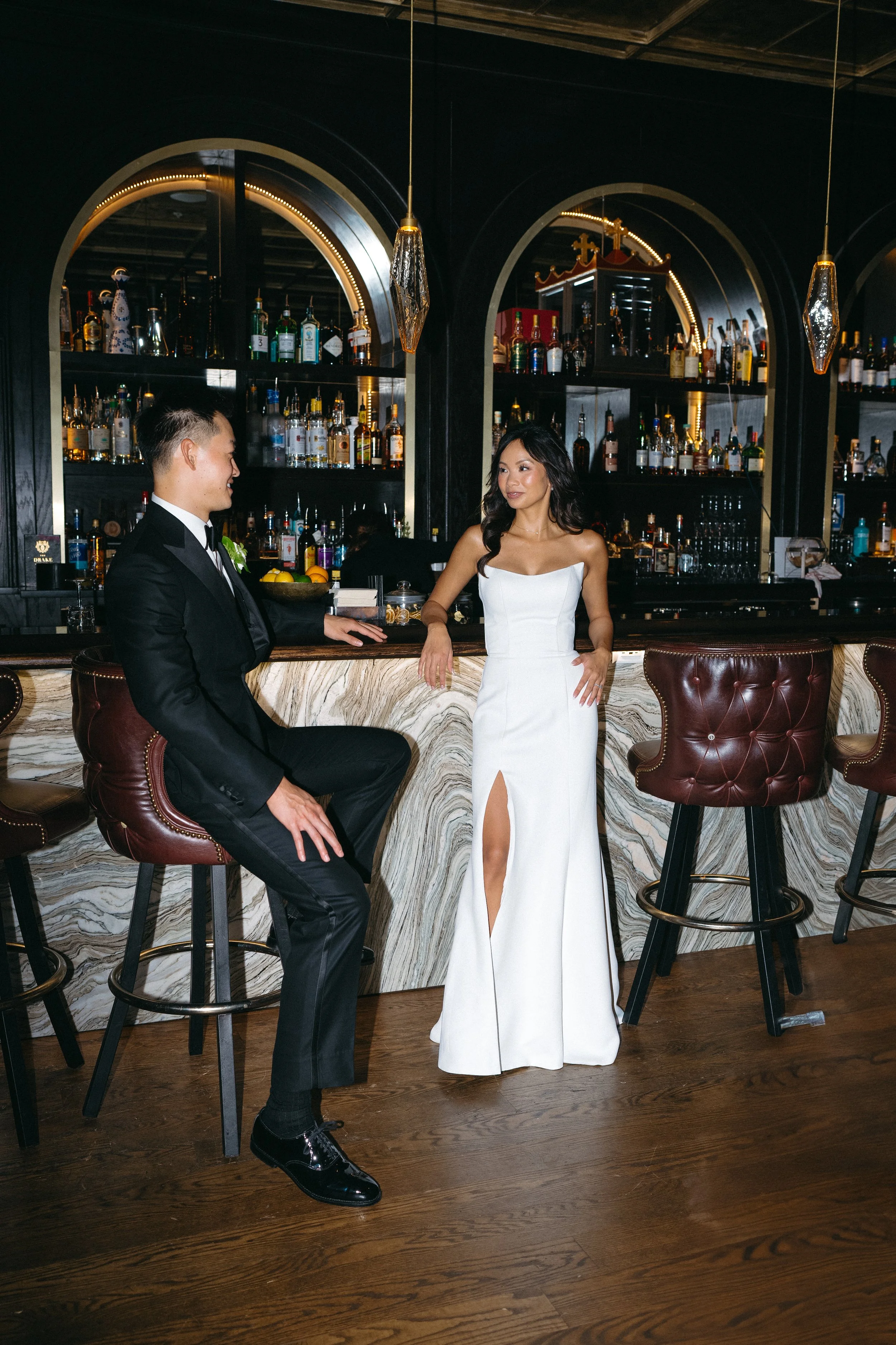 A man in a black tuxedo and a woman in a white dress are standing at a bar, engaged in conversation. The bar has a dark background with shelves of various liquor bottles and modern light fixtures hanging from the ceiling.