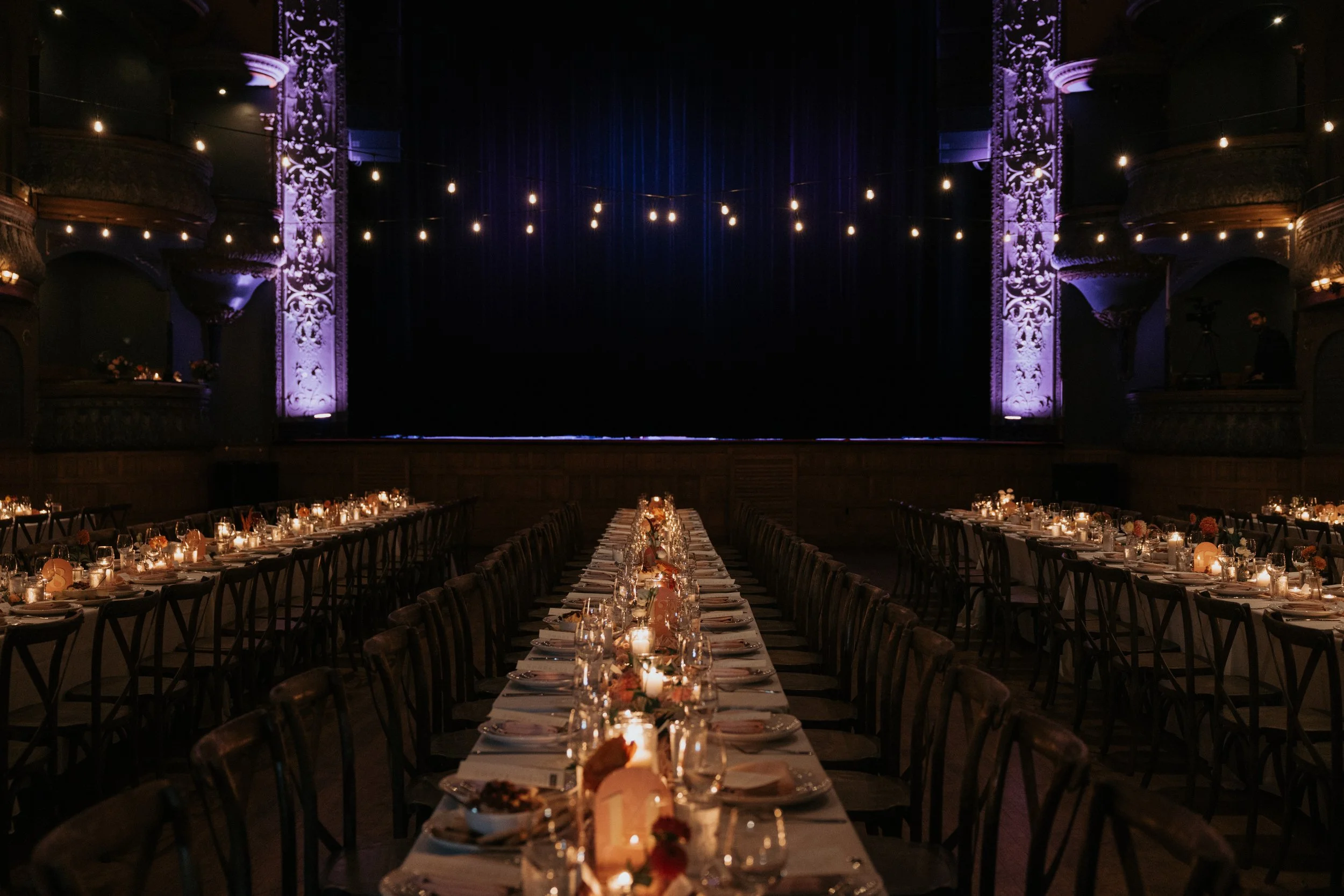 A dimly lit banquet hall set up for a formal event with a long dining table, candles, flowers, and overhead string lights, with an ornate stage or backdrop in the background.