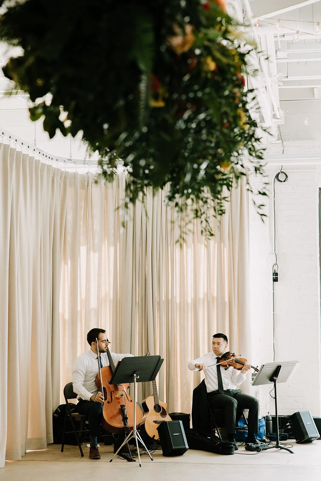 Two men playing string instruments, a cello and a violin, in a room with cream curtains, a white brick wall, and string lights.