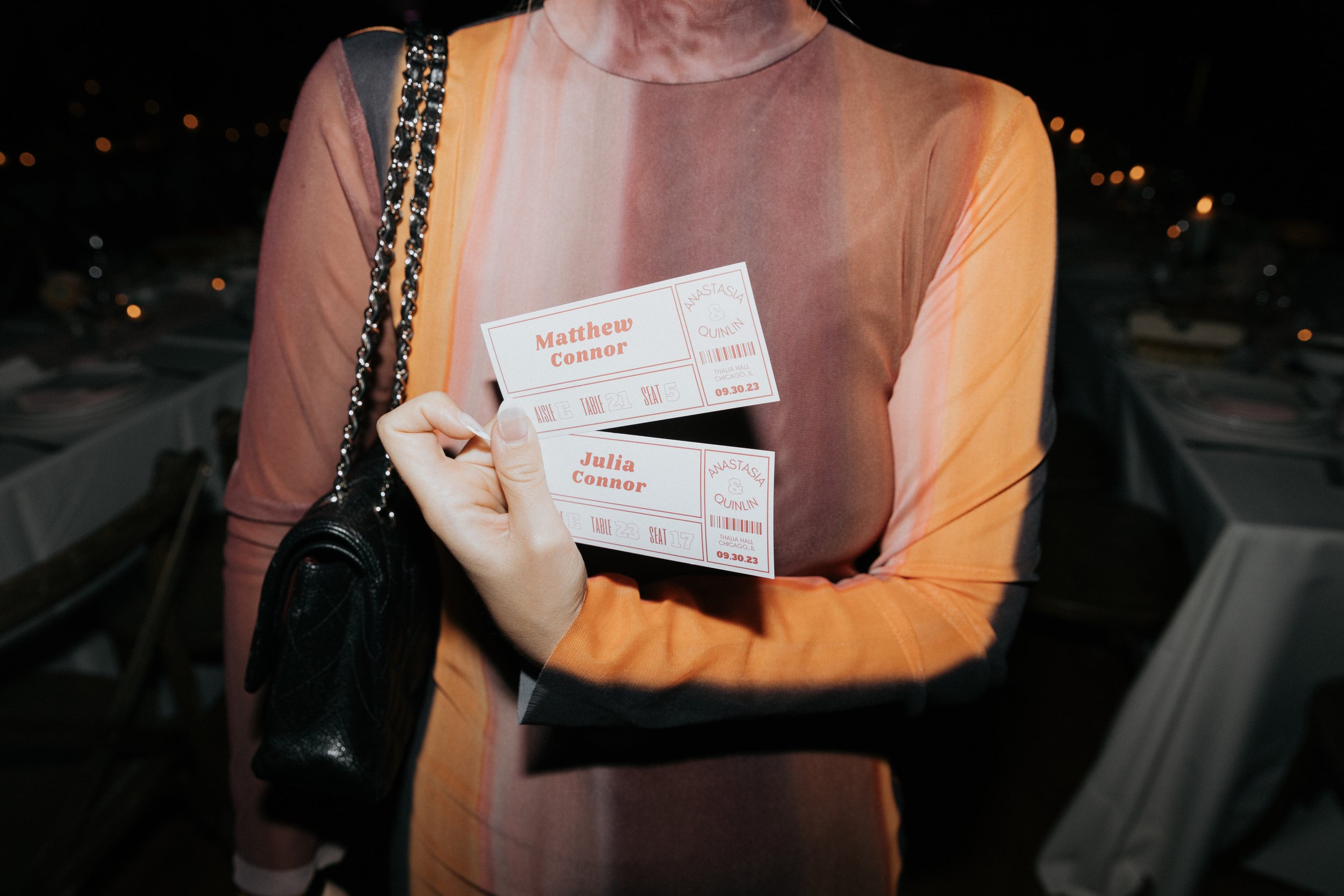 Person holding two event tickets with names Matthew Connor and Julia Connor, against a dark background with scattered lights, wearing an orange and pink top and a black purse with a chain strap.