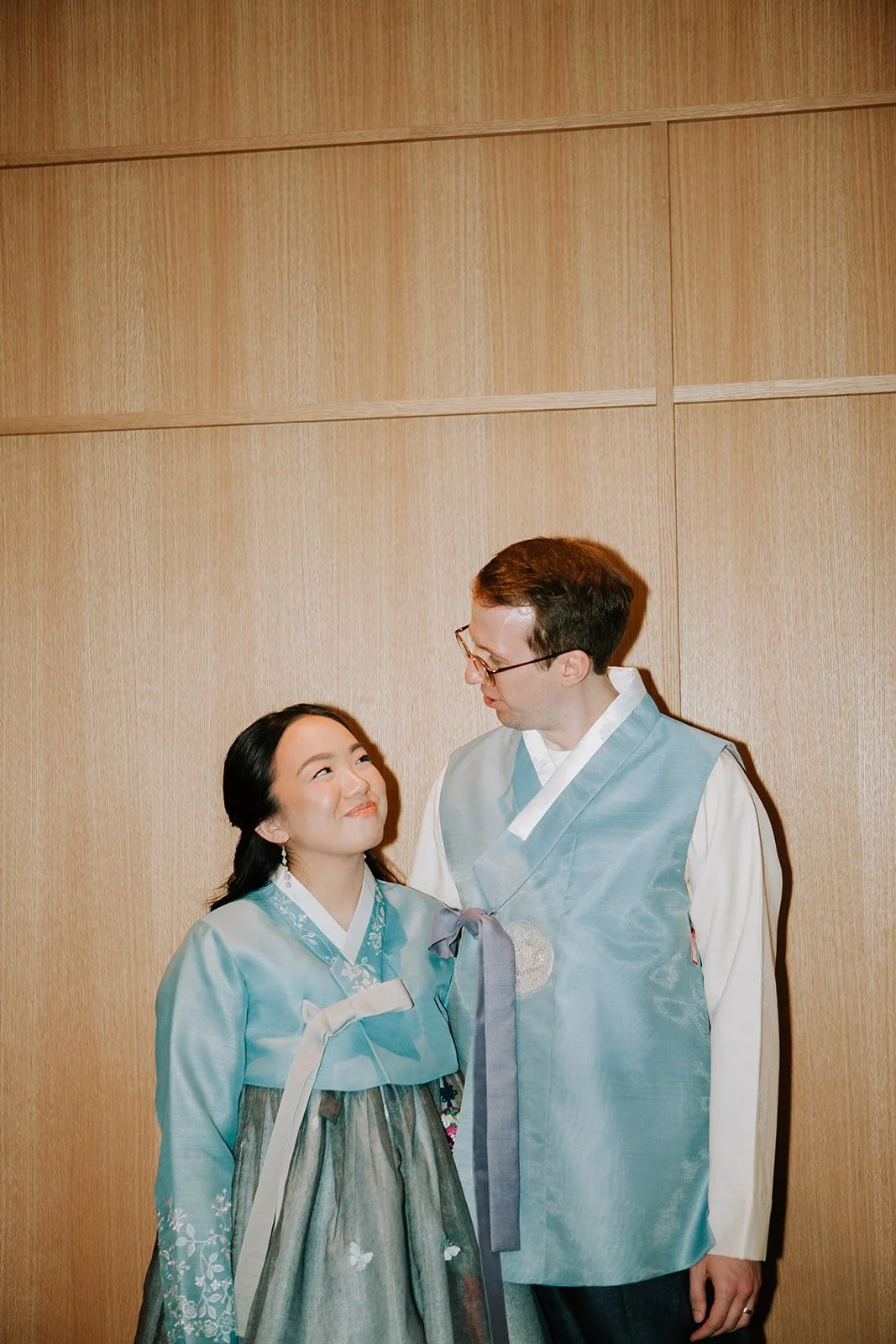 A woman and a man wearing traditional Korean hanbok clothes, standing indoors against a wooden wall, looking at each other and smiling