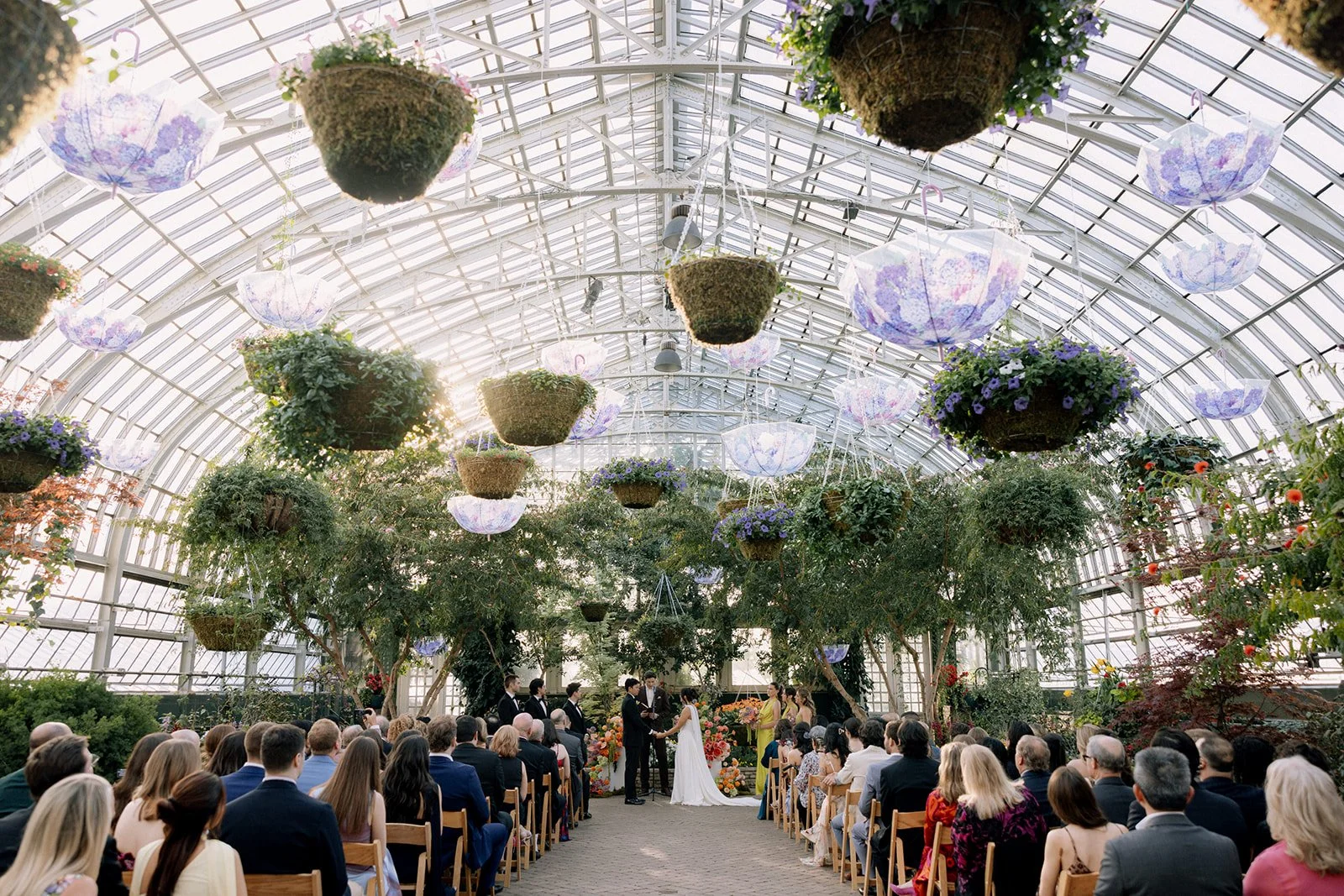 A wedding ceremony inside a greenhouse with hanging flower umbrellas and potted plants. The bride and groom exchange vows in front of an officiant, surrounded by seated guests.