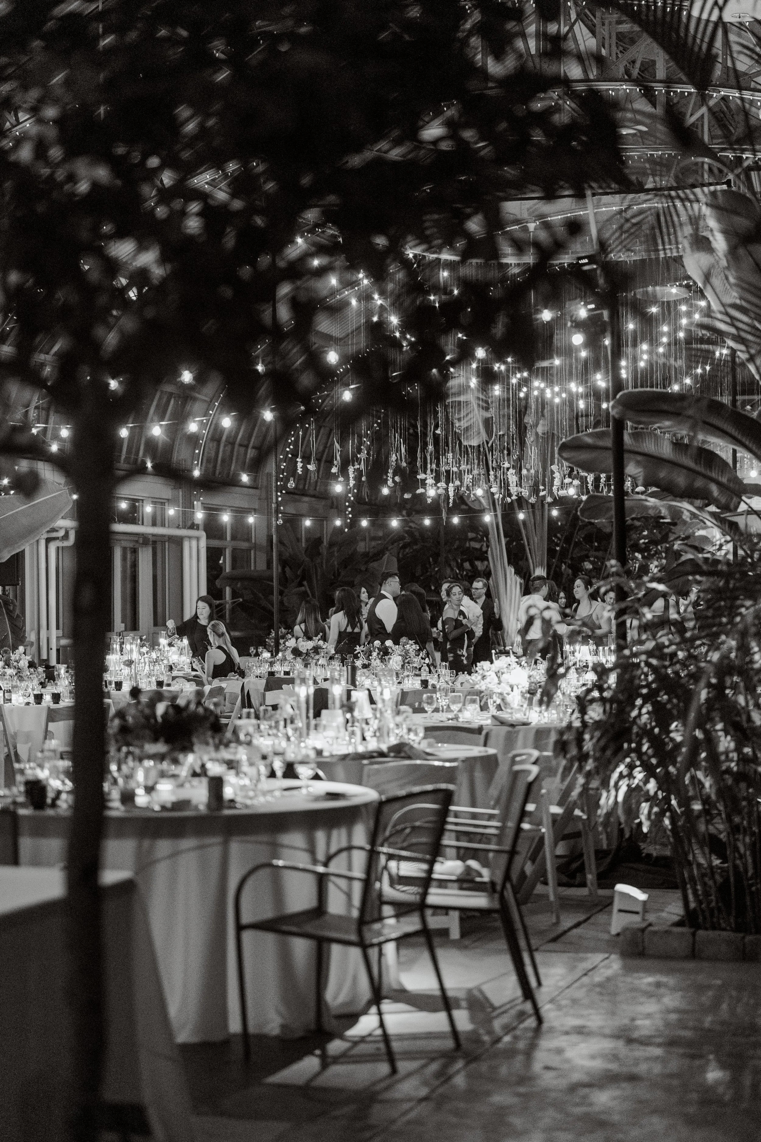 A nighttime indoor event with decorated tables, string lights, and people dressed in formal attire, likely at a wedding or celebration.