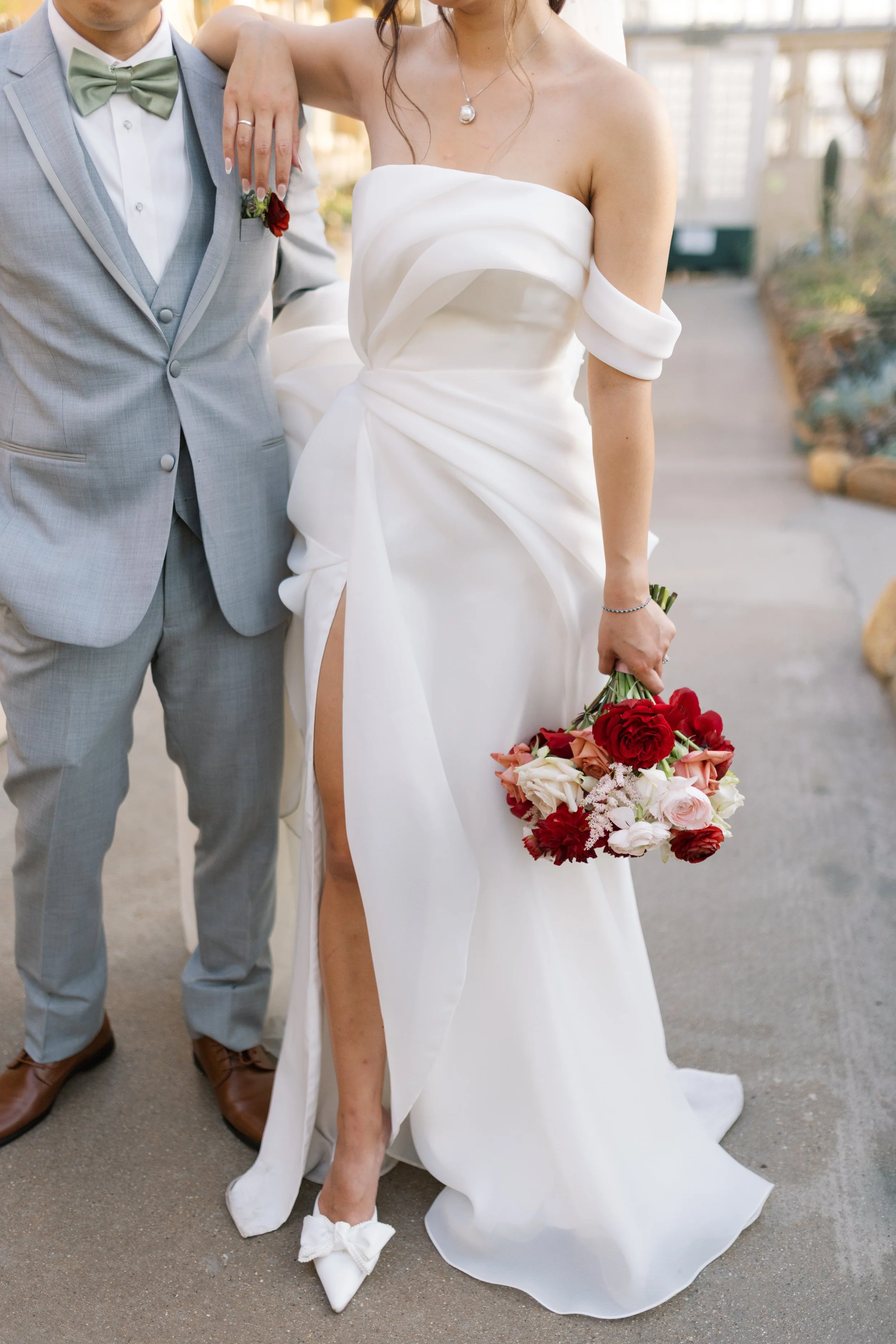 Bride in a white wedding dress holding a bouquet of red and pink flowers standing next to groom in a light gray suit with a green bow tie, outside.