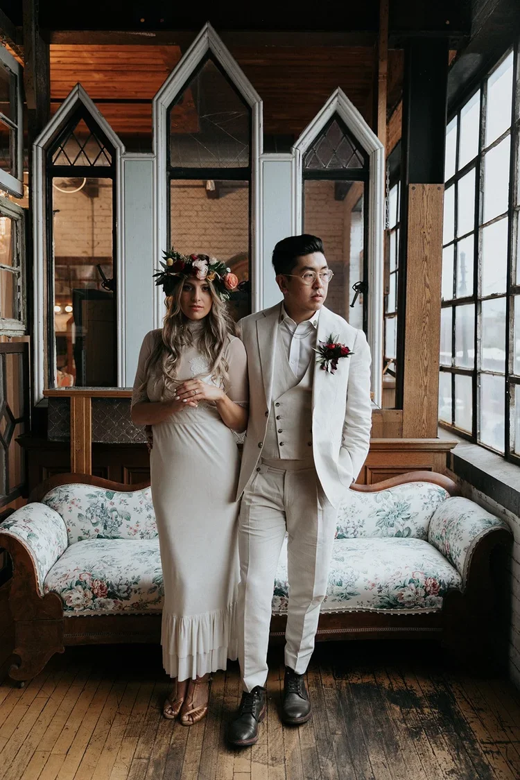 A couple dressed in formal wedding attire standing indoors near a floral-patterned vintage sofa, with large windows to the right and a partition with pointed arches behind them. The woman wears a white dress and a floral crown, while the man wears a 