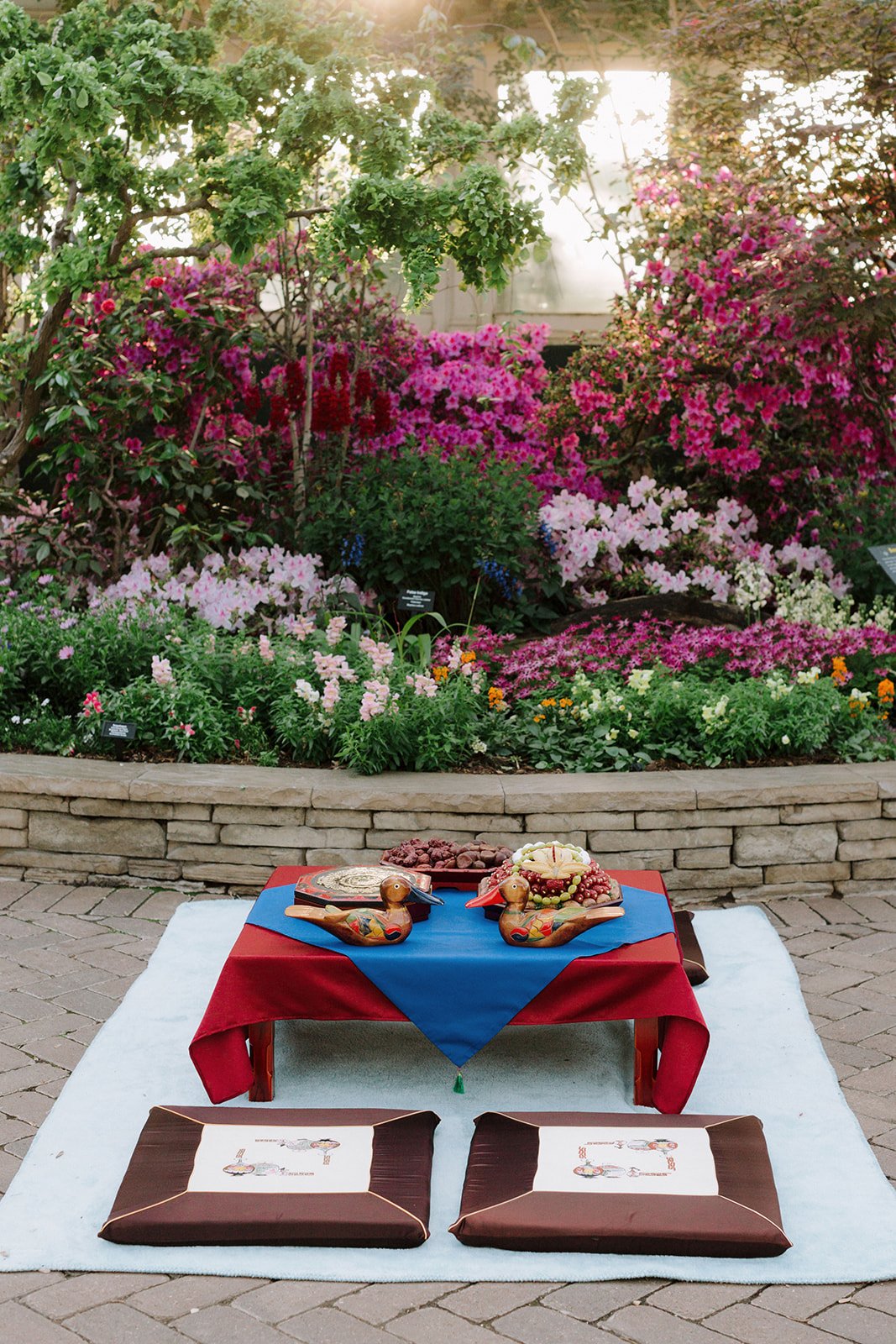 A table set for a traditional Asian meal with two wooden duck-shaped decorations, plates of food, and a blue and red tablecloth, situated on a white rug in front of a vibrant garden with pink, purple, and white flowers.