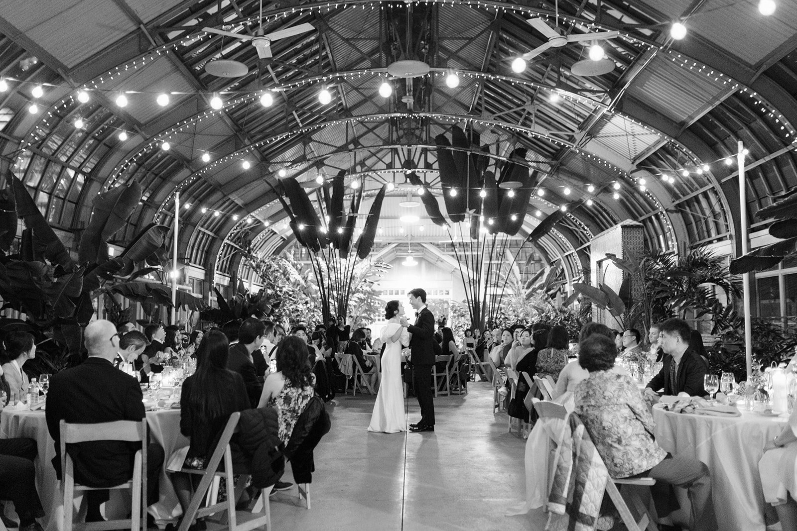 A black and white photo of a wedding reception inside a large, arched greenhouse with string lights. The bride and groom are dancing in the center, surrounded by seated guests at round tables.