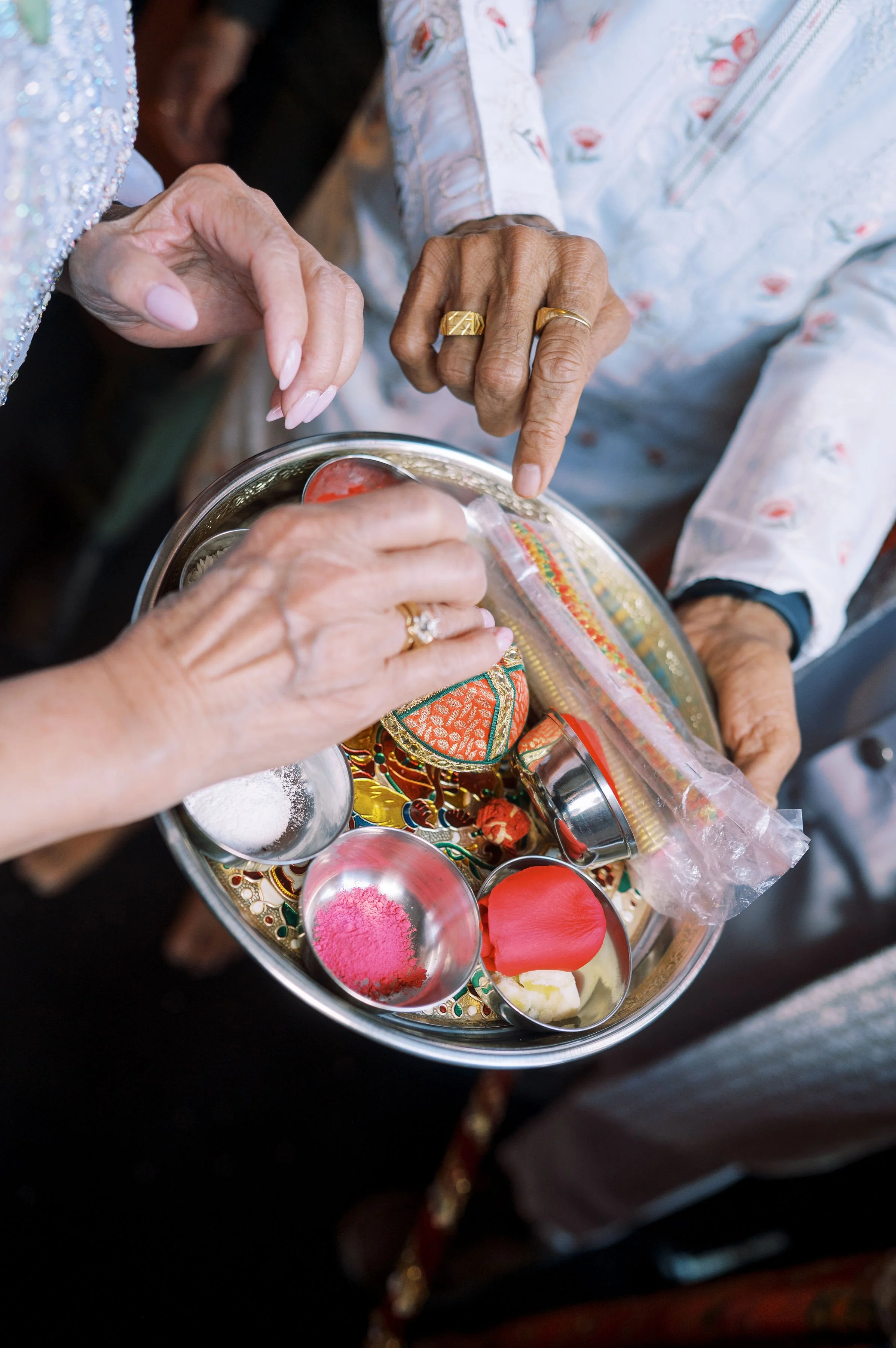 Close-up of elderly hands holding a silver tray with colorful items, including pink, yellow, and red powders, and traditional decorative objects in an outdoor setting.