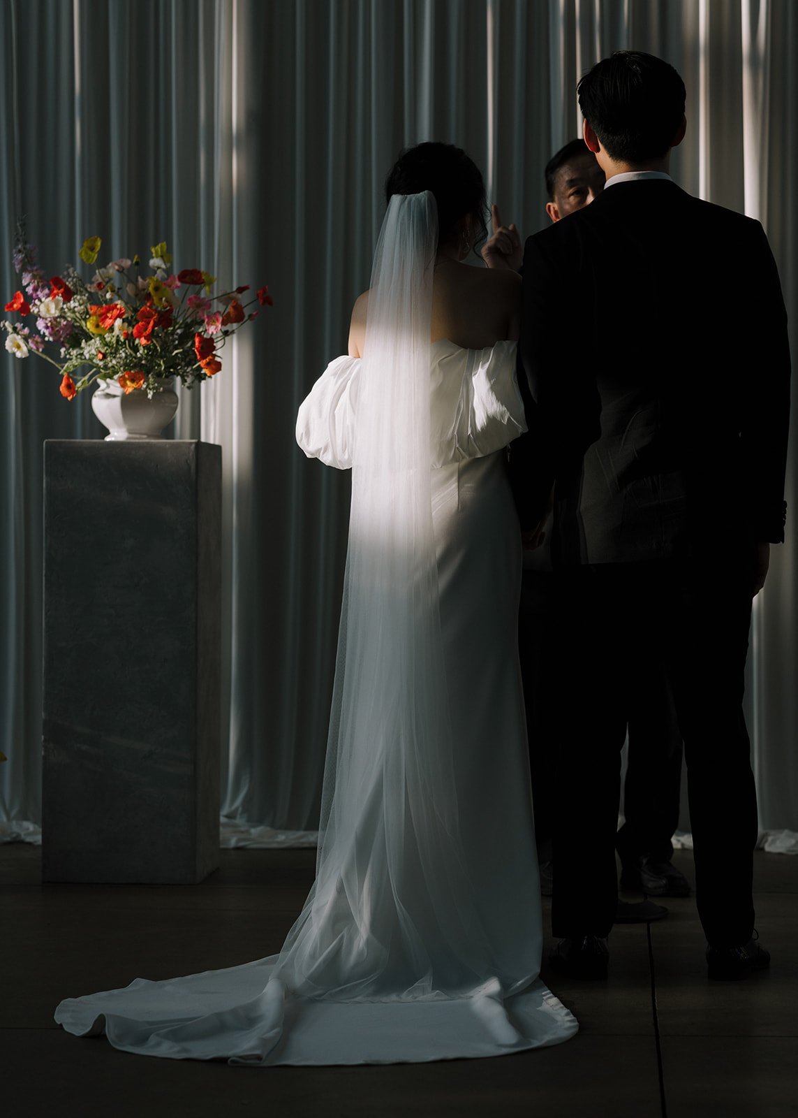 A bride and groom exchanging vows during a wedding ceremony, with a flower arrangement on a pedestal to the side and curtains in the background.