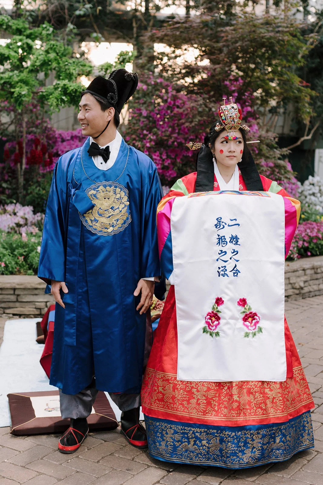 A man and woman dressed in traditional Chinese wedding clothing, standing outdoors with blooming flowers and greenery in the background.
