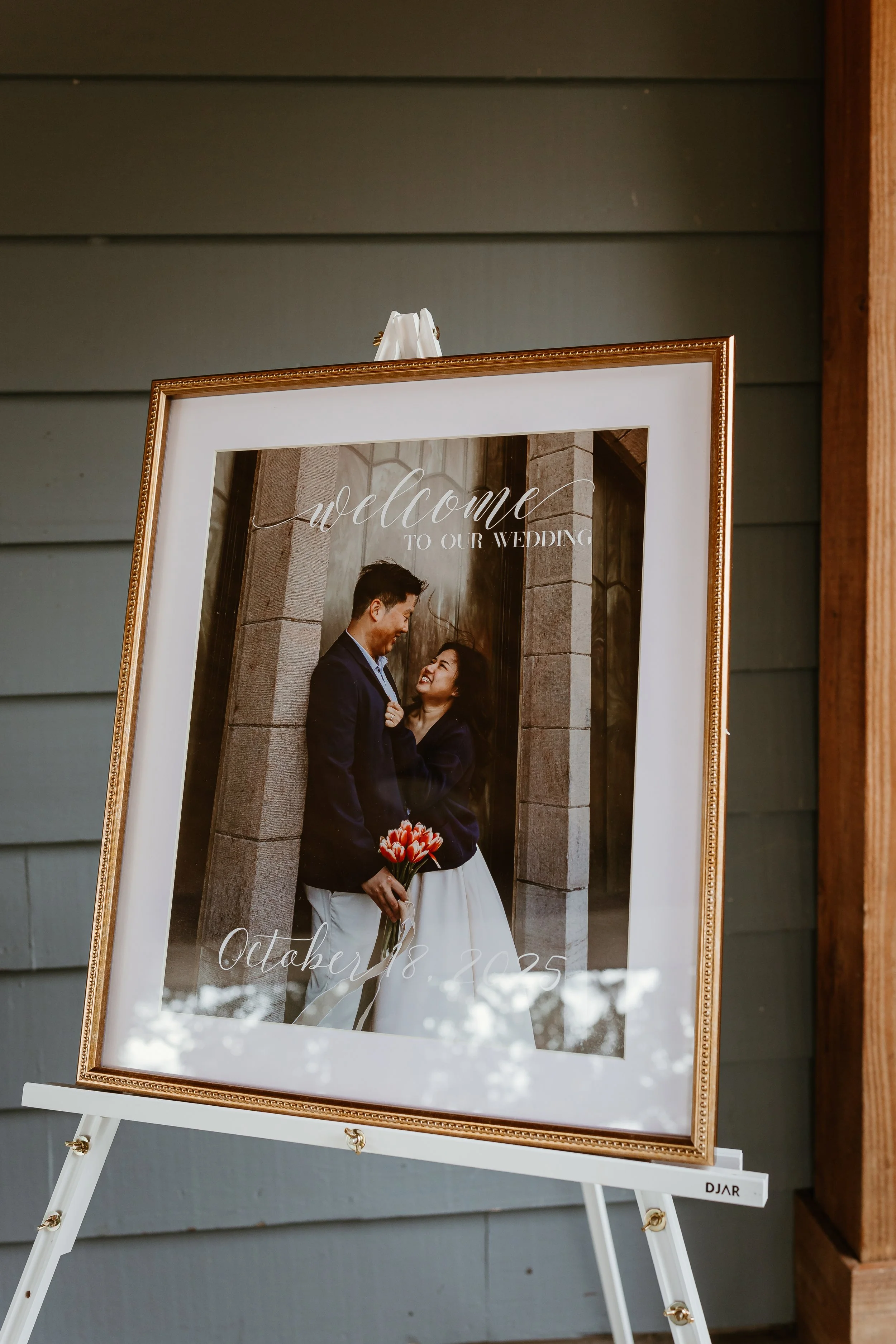 Wedding photo displayed on a stand with a wedding date, showing a couple in wedding attire looking at each other, with one woman holding a bouquet of pink tulips, in front of a gray wall.