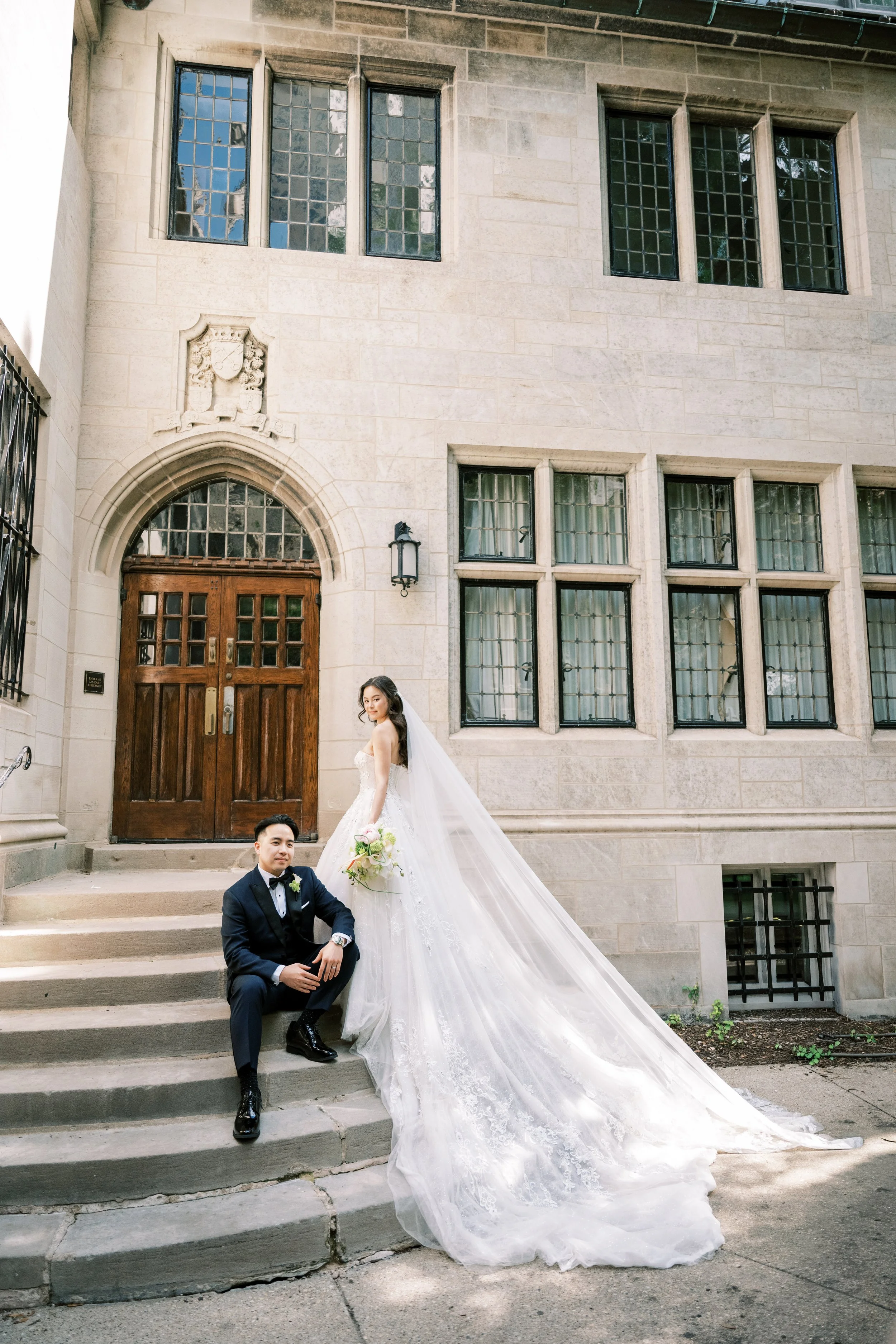 A bride and groom pose on stone steps in front of a historic stone building with large windowpanes and a wooden door. The bride, in a white wedding gown with a long train and veil, stands on the steps, while the groom, in a black tuxedo, is seated on