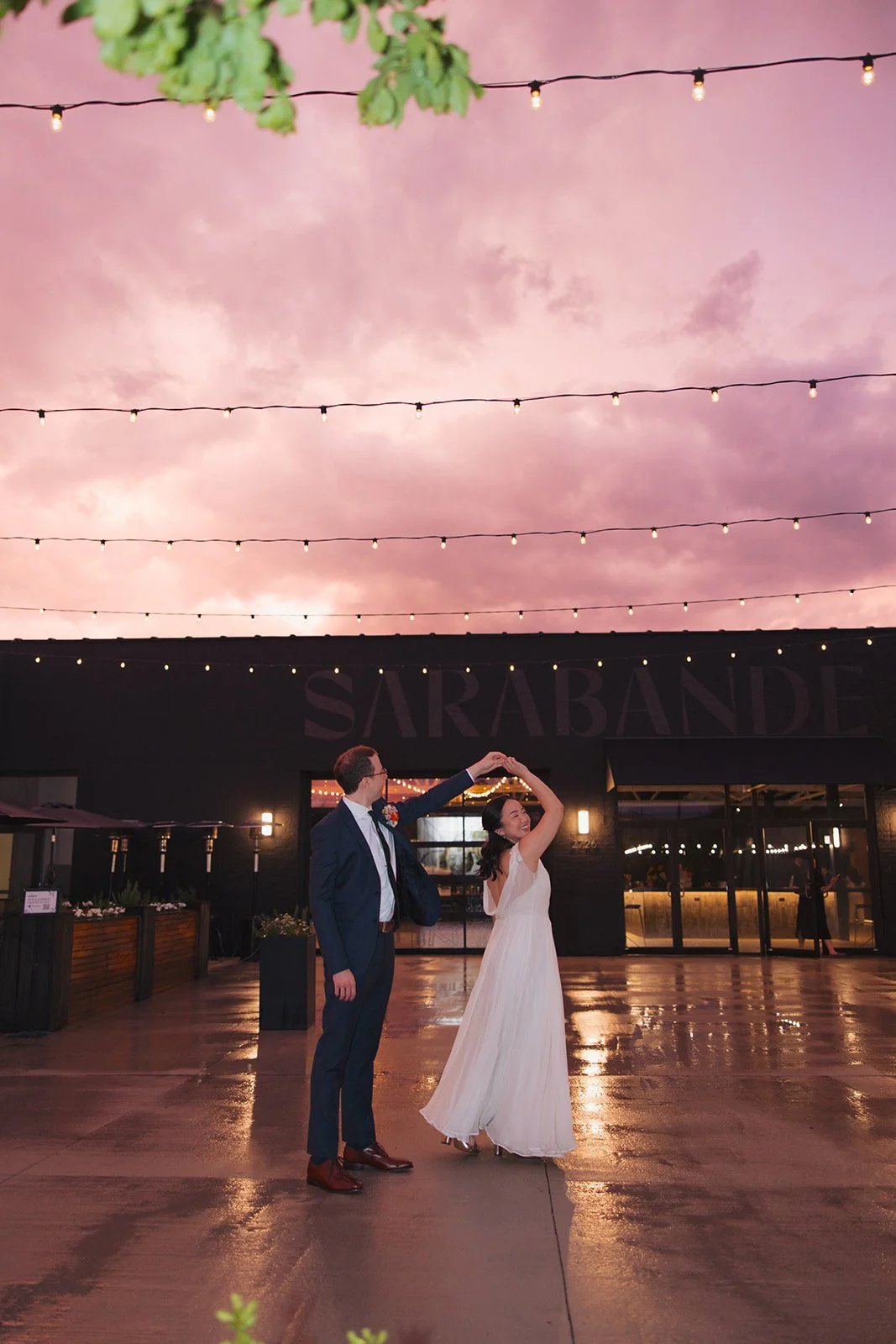 A couple dancing outside during sunset under string lights, with the woman in a white dress and the man in a navy suit, in front of a building with the word 'SARBANDE' on it.