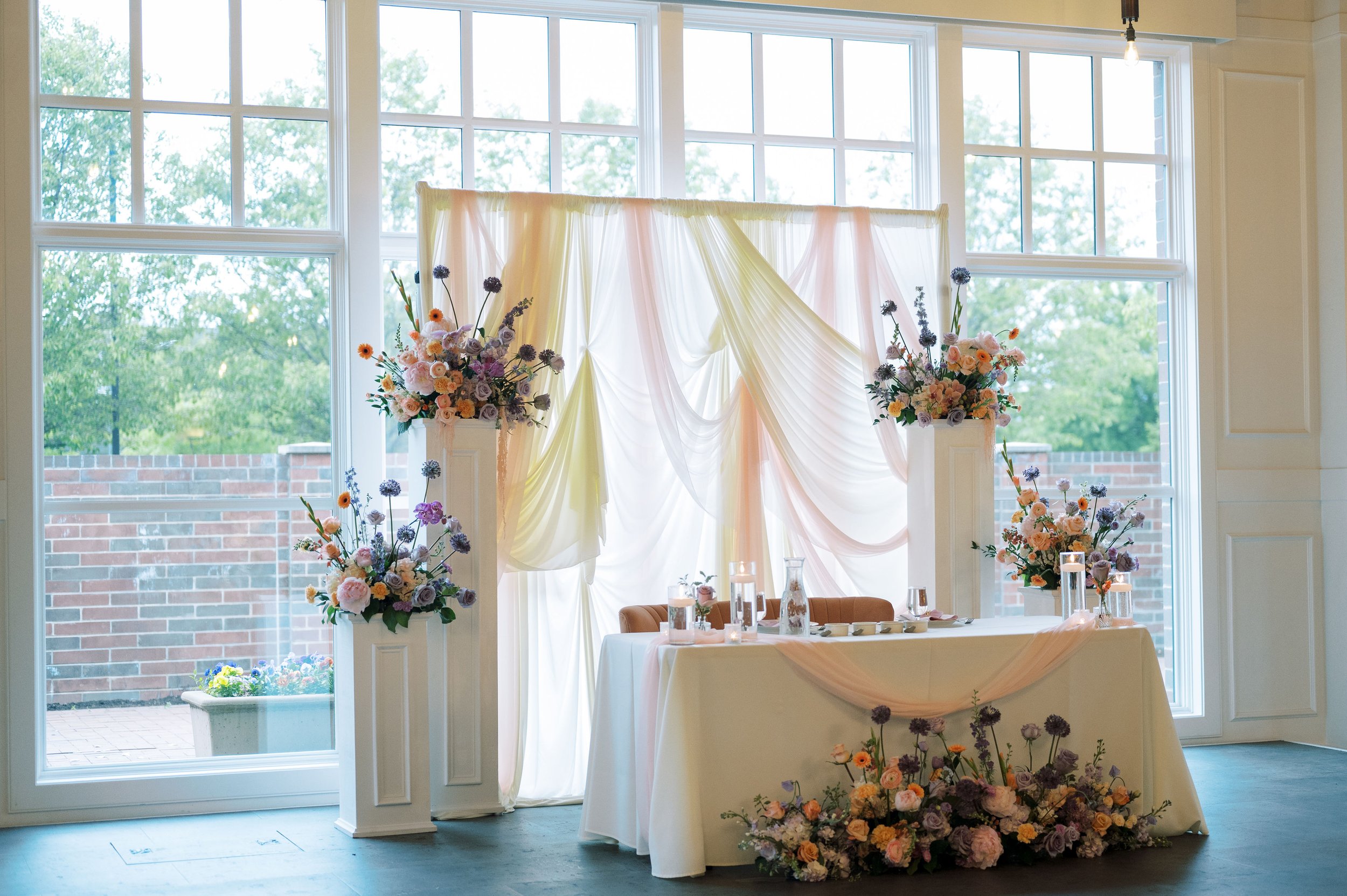 Elegant wedding floral arrangement with pastel-colored flowers and draped fabric backdrop inside a room with large windows and greenery outside.