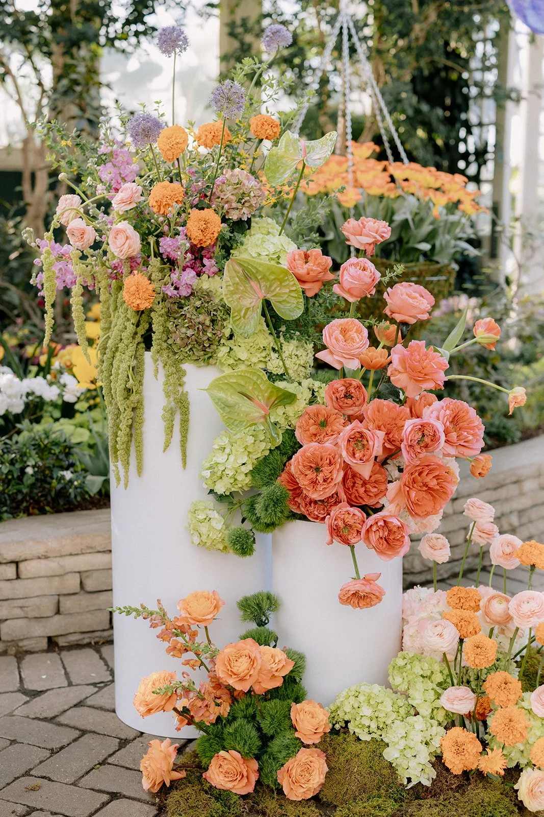 Arrangement of vibrant orange and pink flowers, green foliage, and hanging plants in white cylindrical containers outdoors.