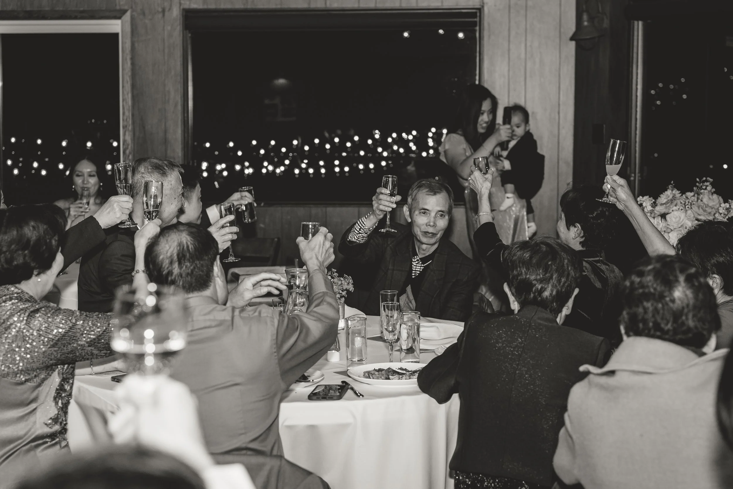 A group of people celebrating at a banquet table, raising glasses in a toast, with a woman in the background taking a photo of a young child.