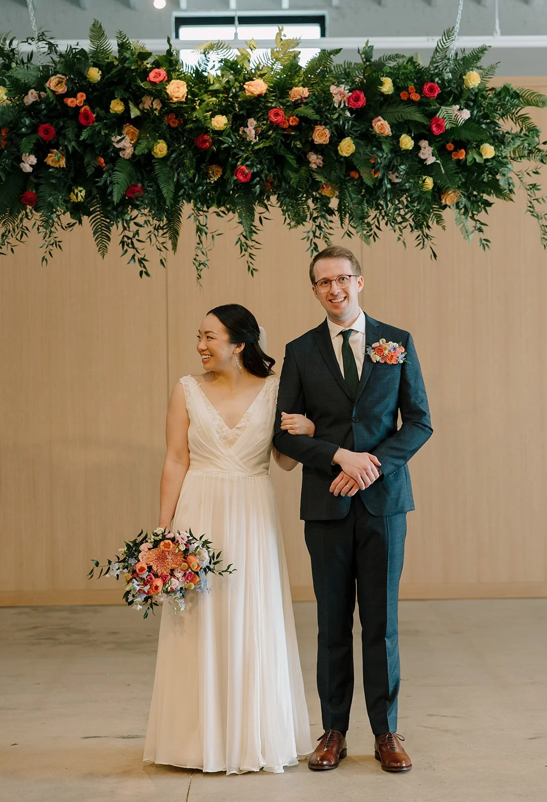 A happy bride and groom standing together indoors under a large floral arrangement of mixed flowers and greenery.